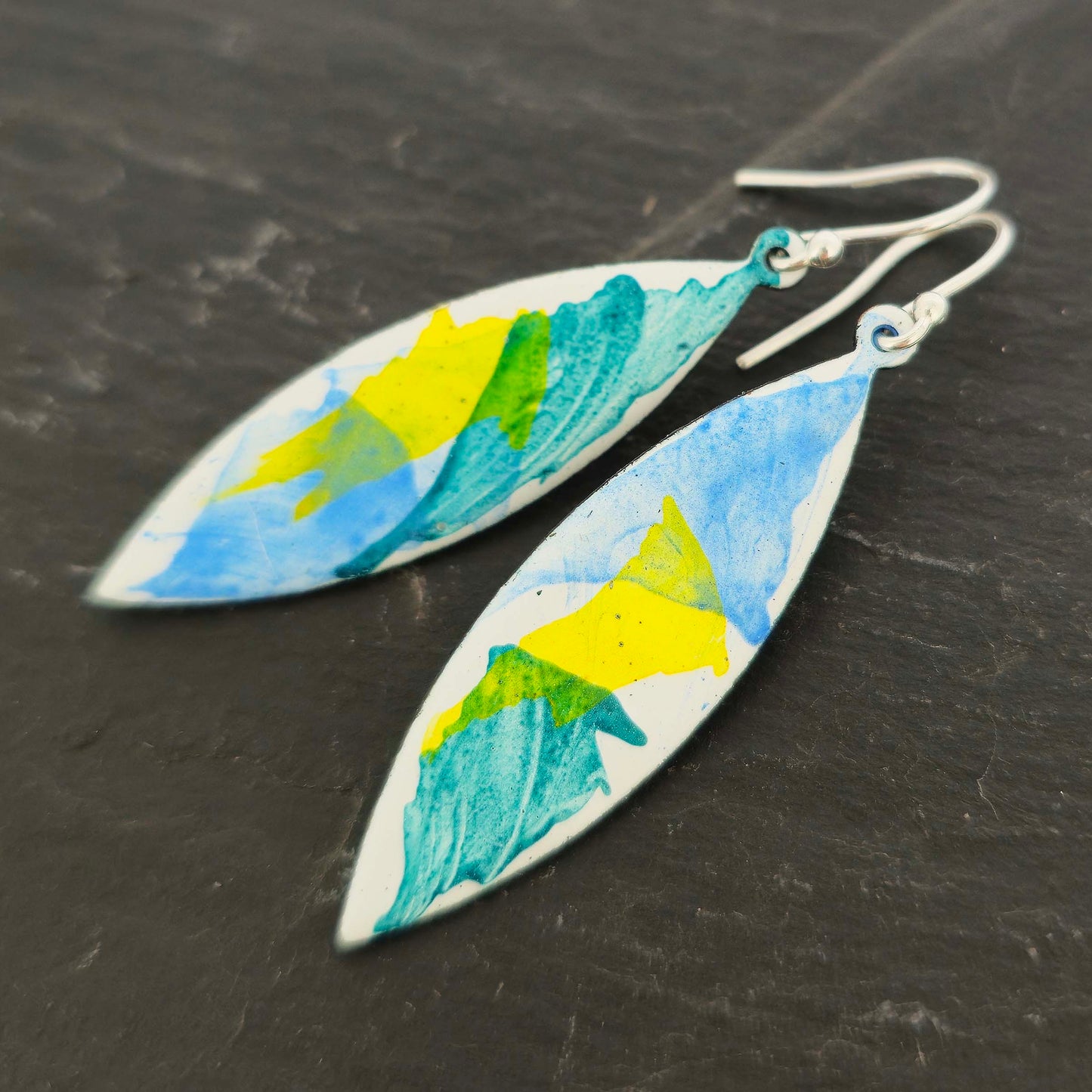 Navette shaped enamel drop earrings with green, blue and yellow splashes on a white background. Shown on slate.