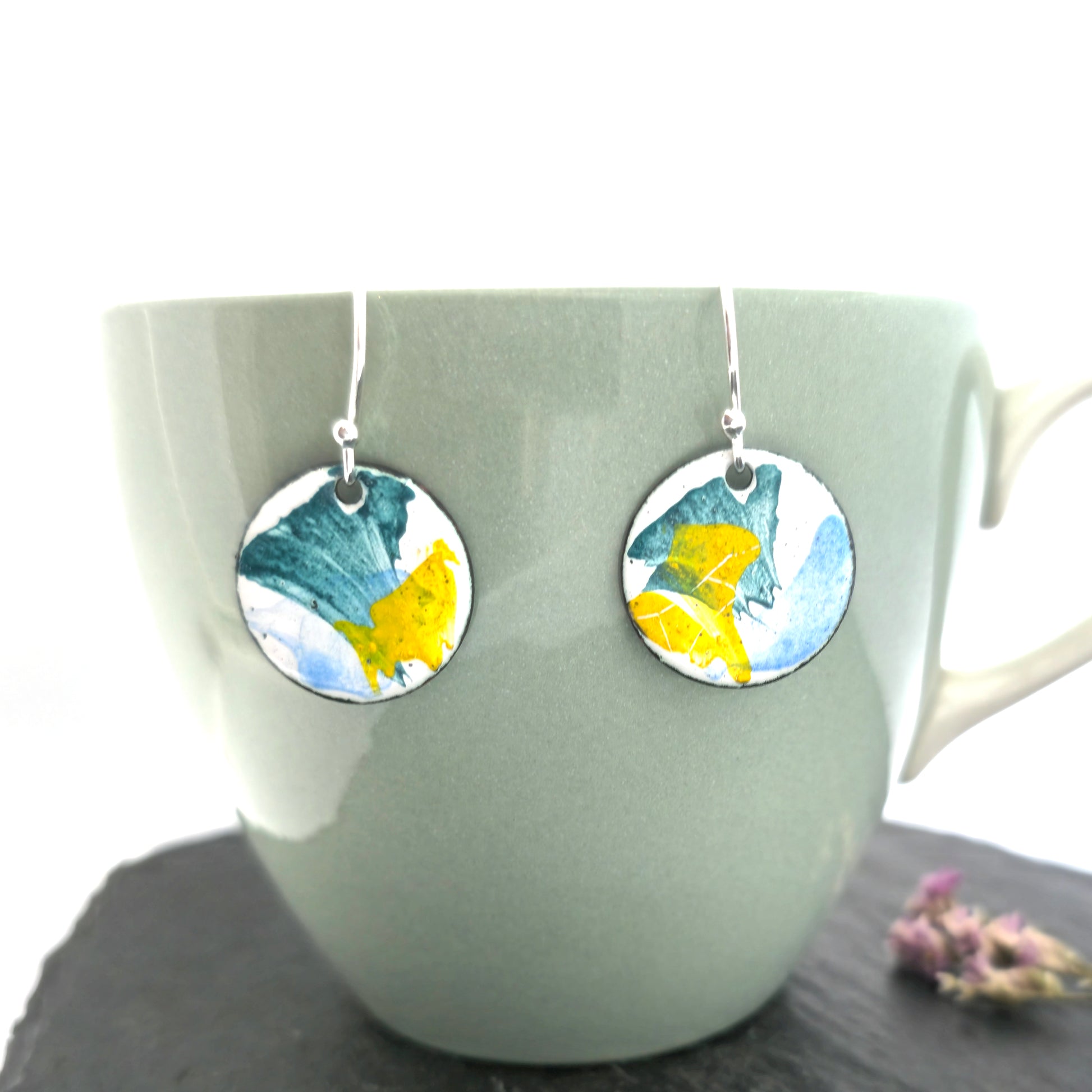 Round enamel drop earrings with splashes of blue, green and yellow on a white background. On silver ear hooks. Pictured on a cup.