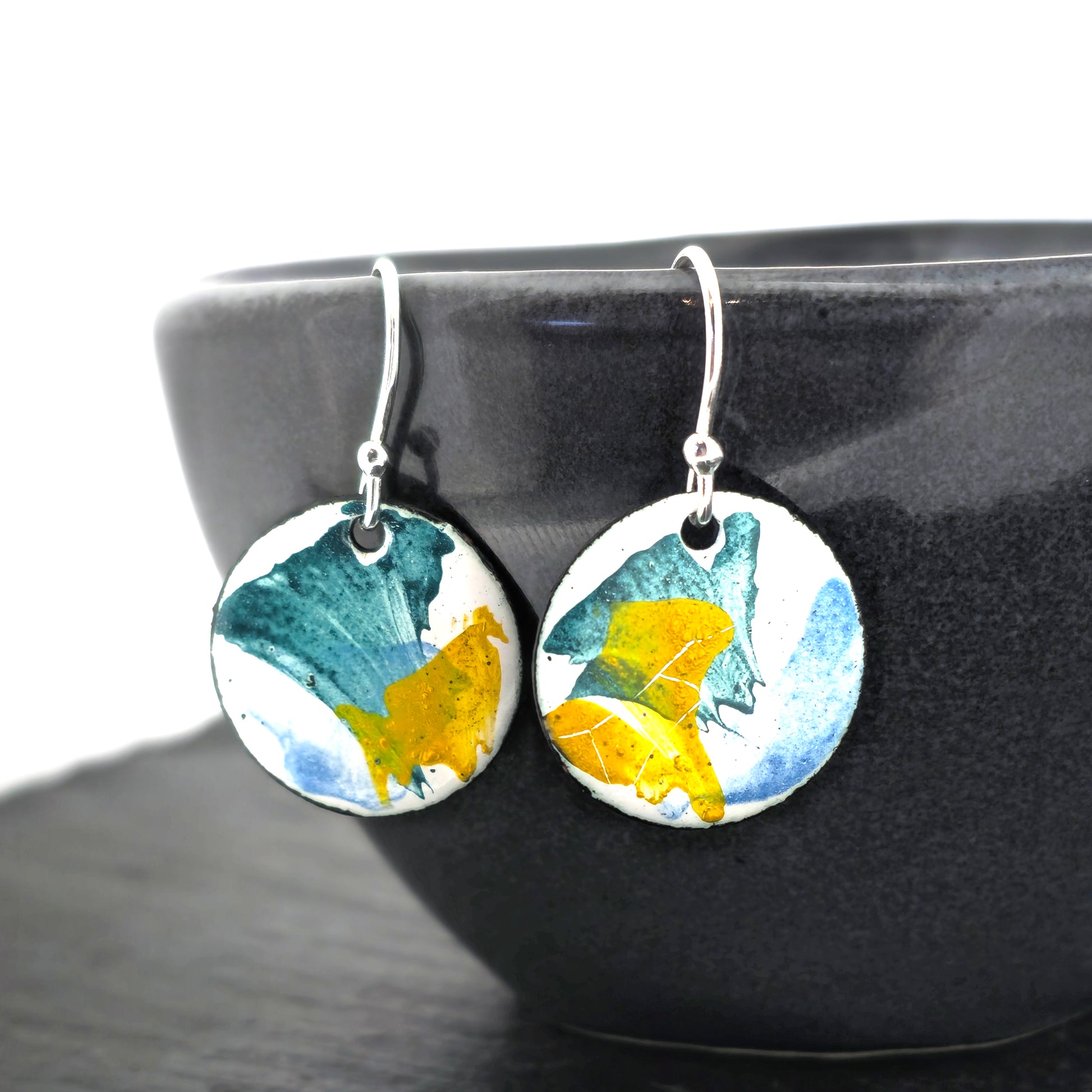 Round enamel drop earrings with splashes of blue, green and yellow on a white background. On silver ear hooks. Pictured on a black bowl.