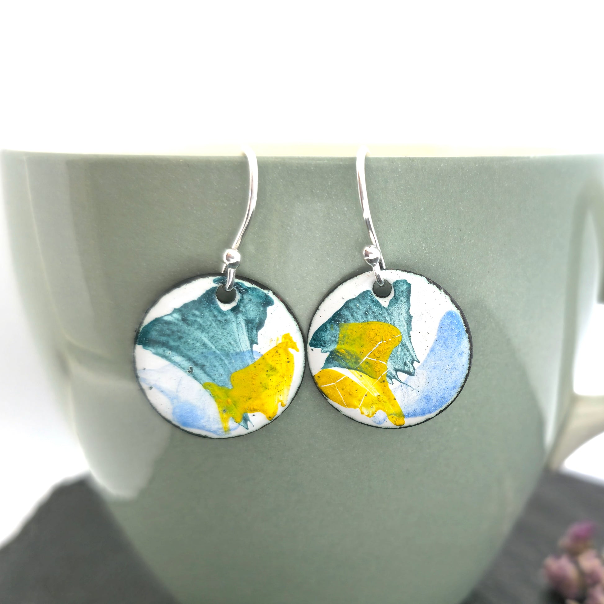 Round enamel drop earrings with splashes of blue, green and yellow on a white background. On silver ear hooks. Pictured on a mug.