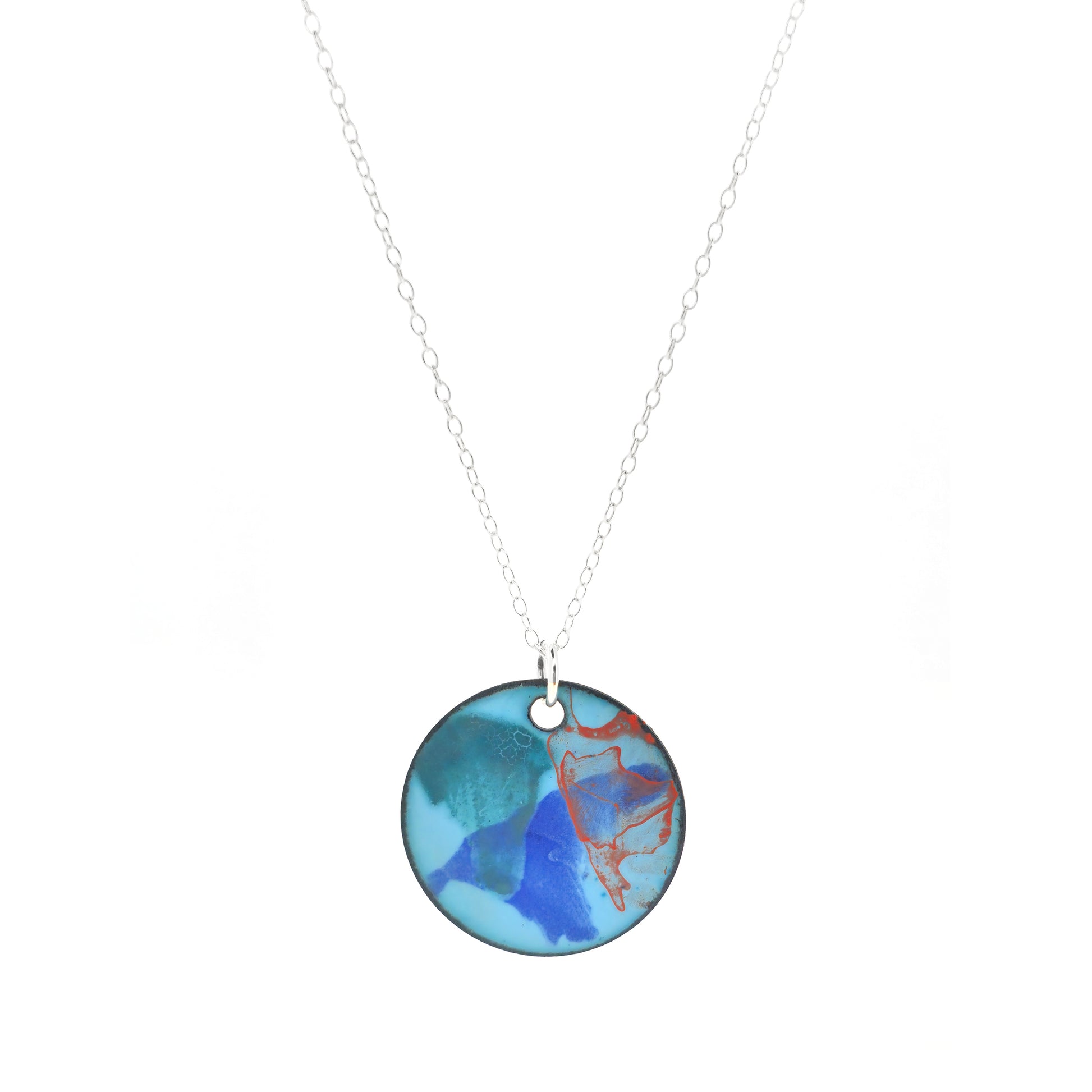 A round enamel pendant with a pale blue background and splashes of green, blue and orange. Suspended from a silver chain.