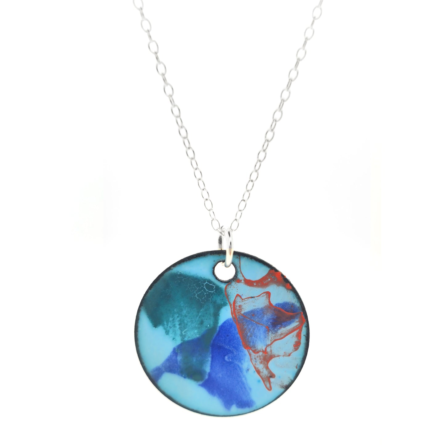 A round enamel pendant with a pale blue background and splashes of green, blue and orange. Suspended from a silver chain.
