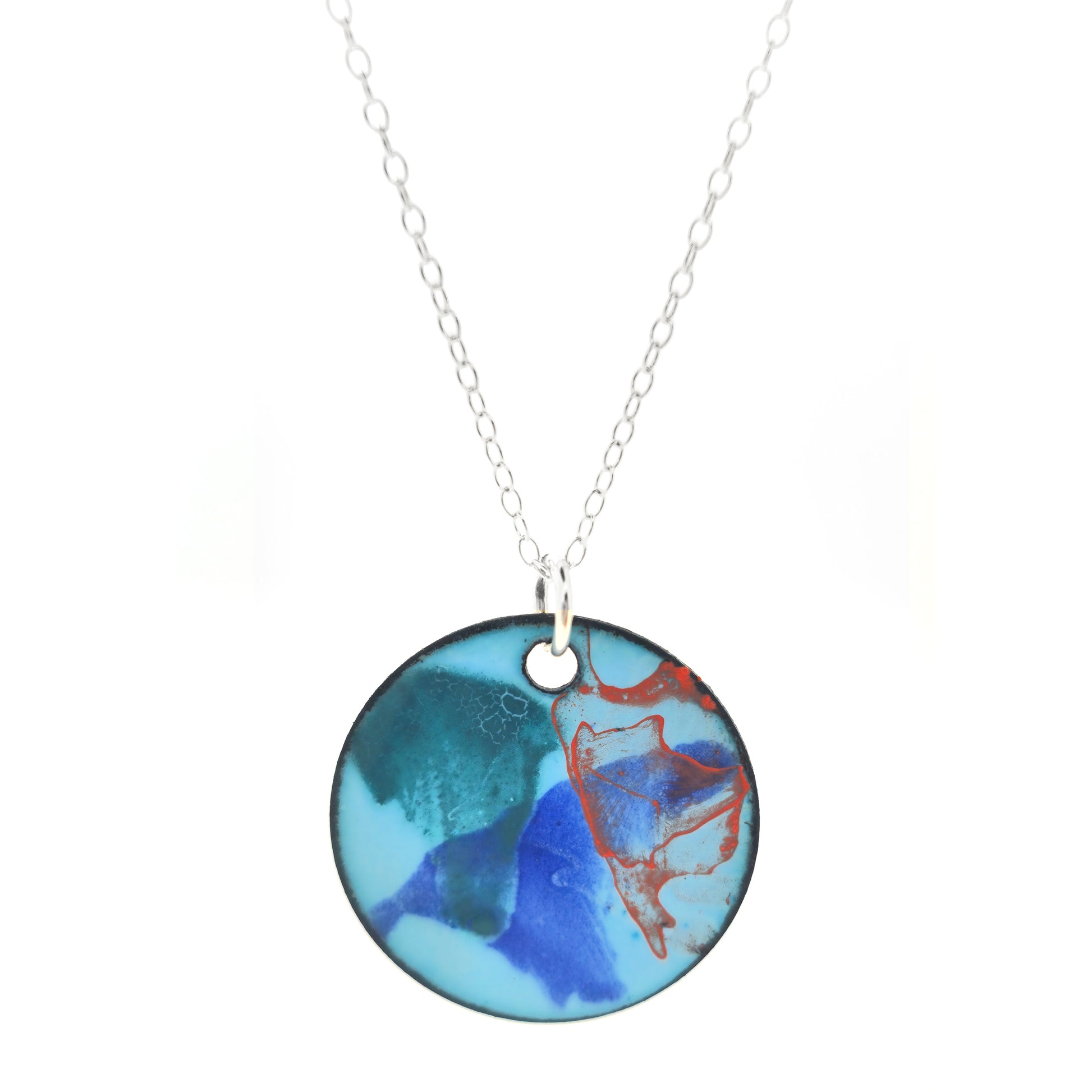 A round enamel pendant with a pale blue background and splashes of green, blue and orange. Suspended from a silver chain.