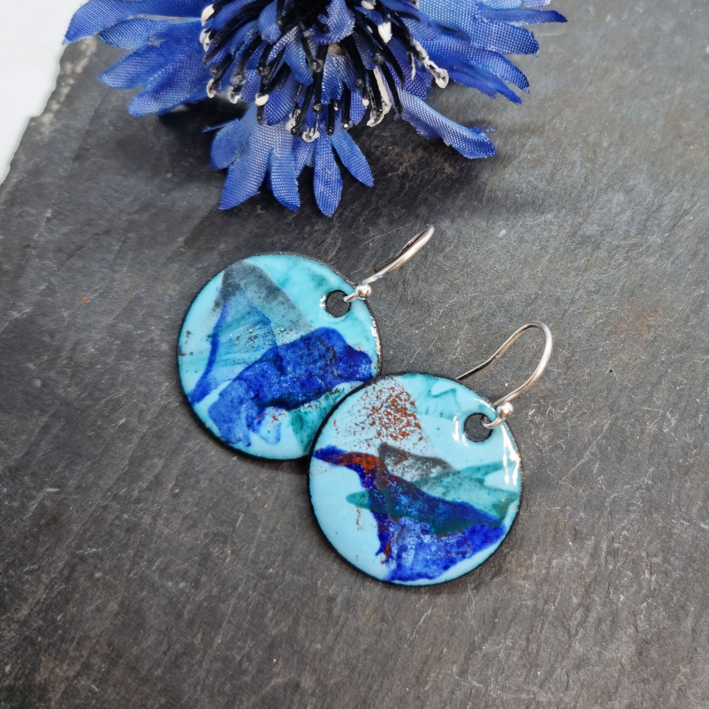 Round enamel drop earrings with splashes of dark blue, grey, green and red on a light blue background with flower