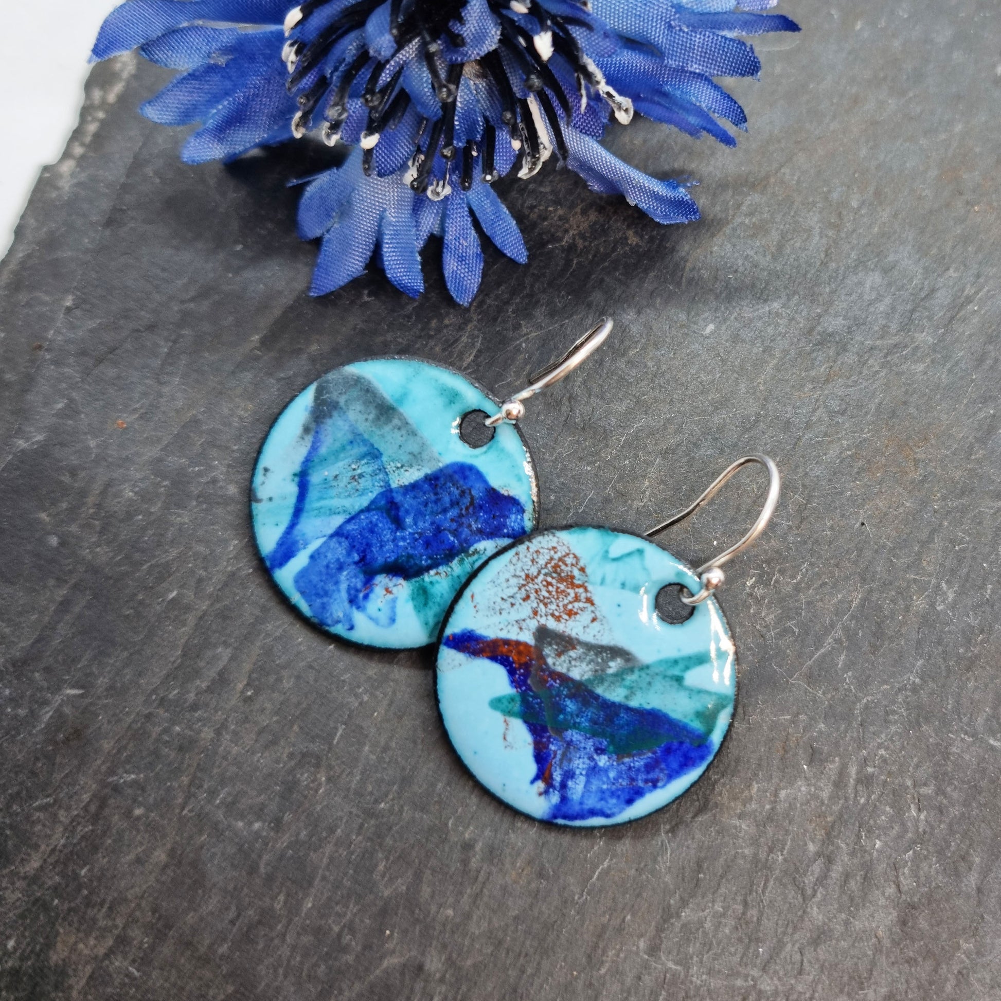 Round enamel drop earrings with splashes of dark blue, grey, green and red on a light blue background with flower