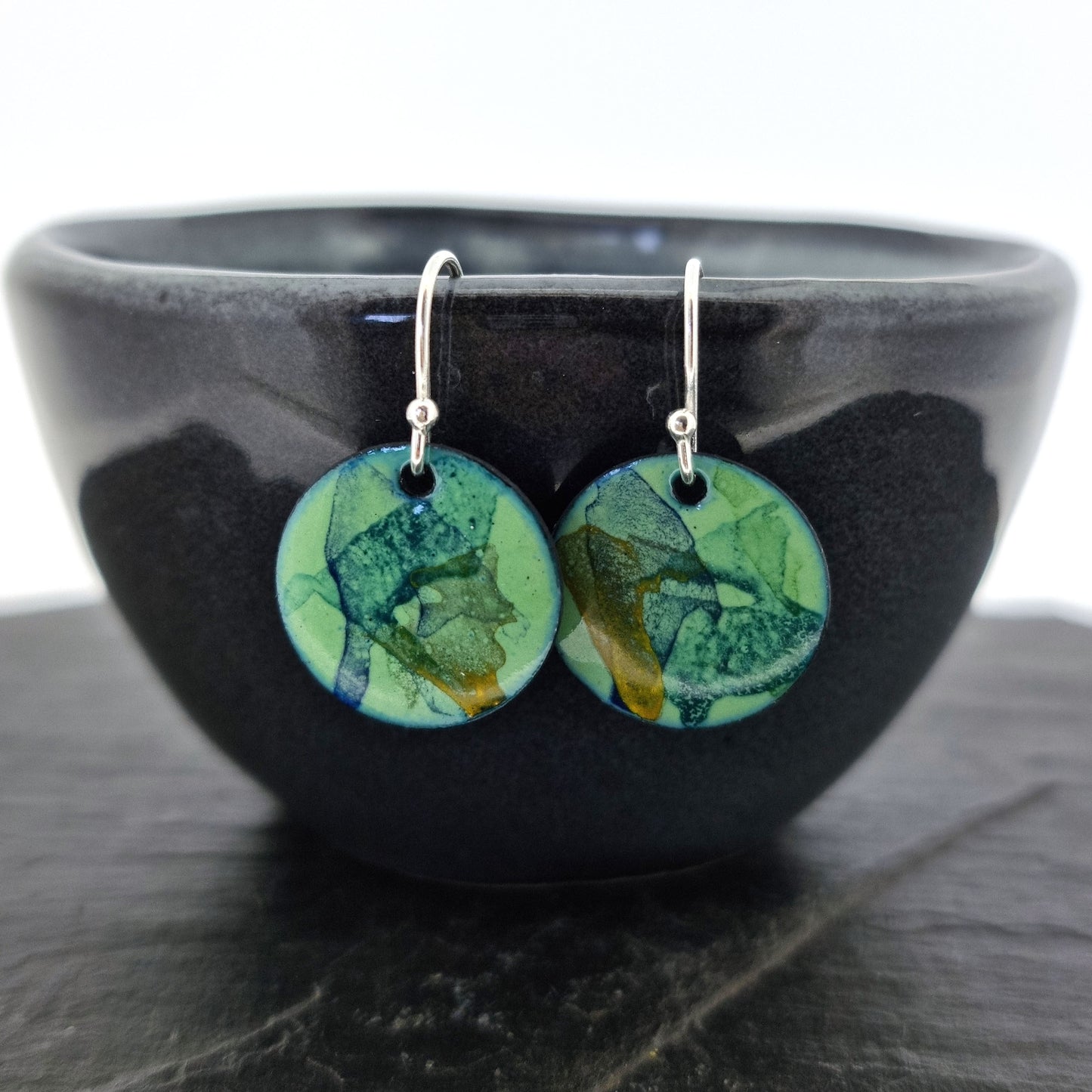 Round enamel drop earrings with splashes of blue, green and yellow on a light green background. Pictured on a bowl.