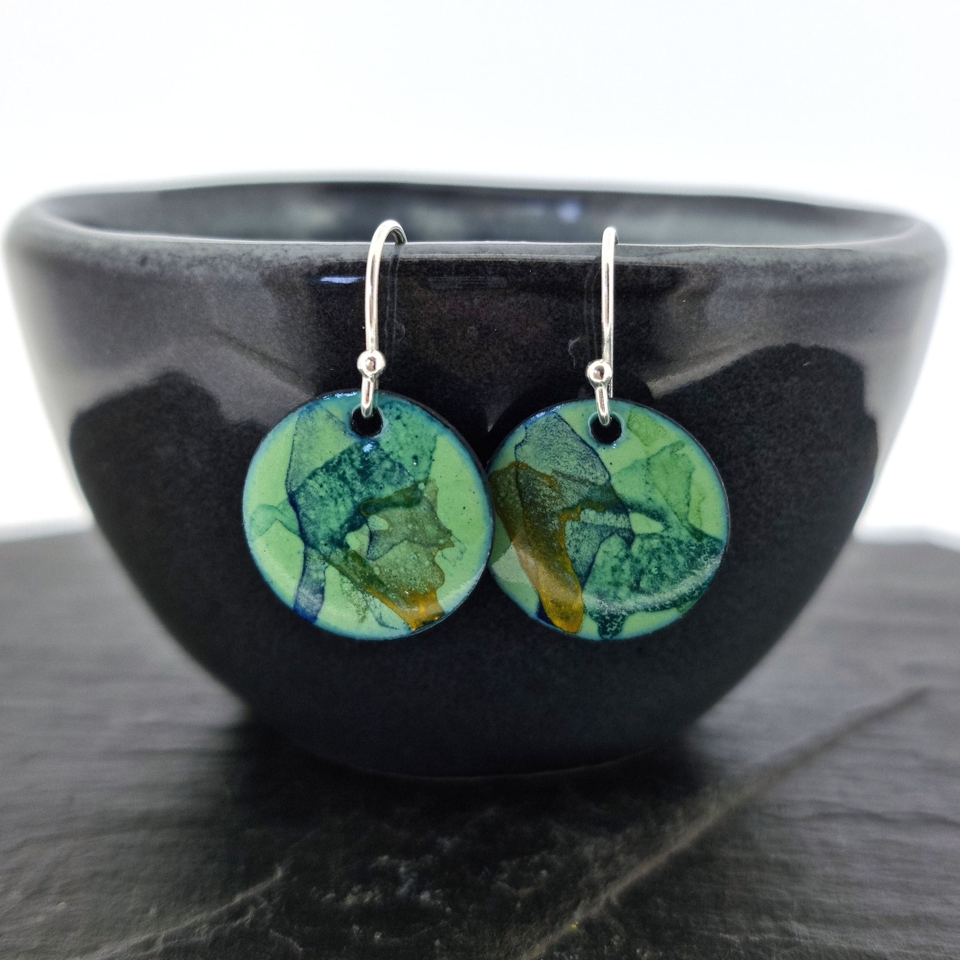 Round enamel drop earrings with splashes of blue, green and yellow on a light green background. Pictured on a bowl.