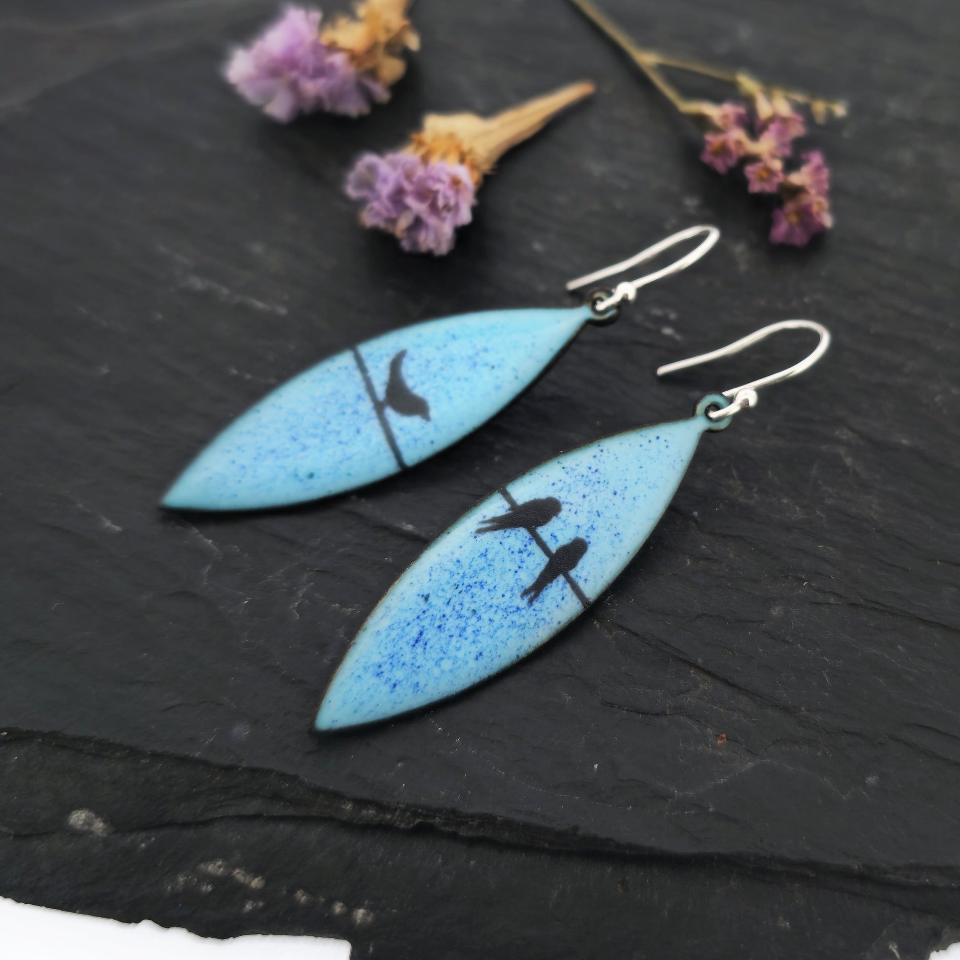Boat-shaped enamel drop earrings with black coloured bird silhouettes sitting on a wire on a blue background. Pictured on slate with flowers.