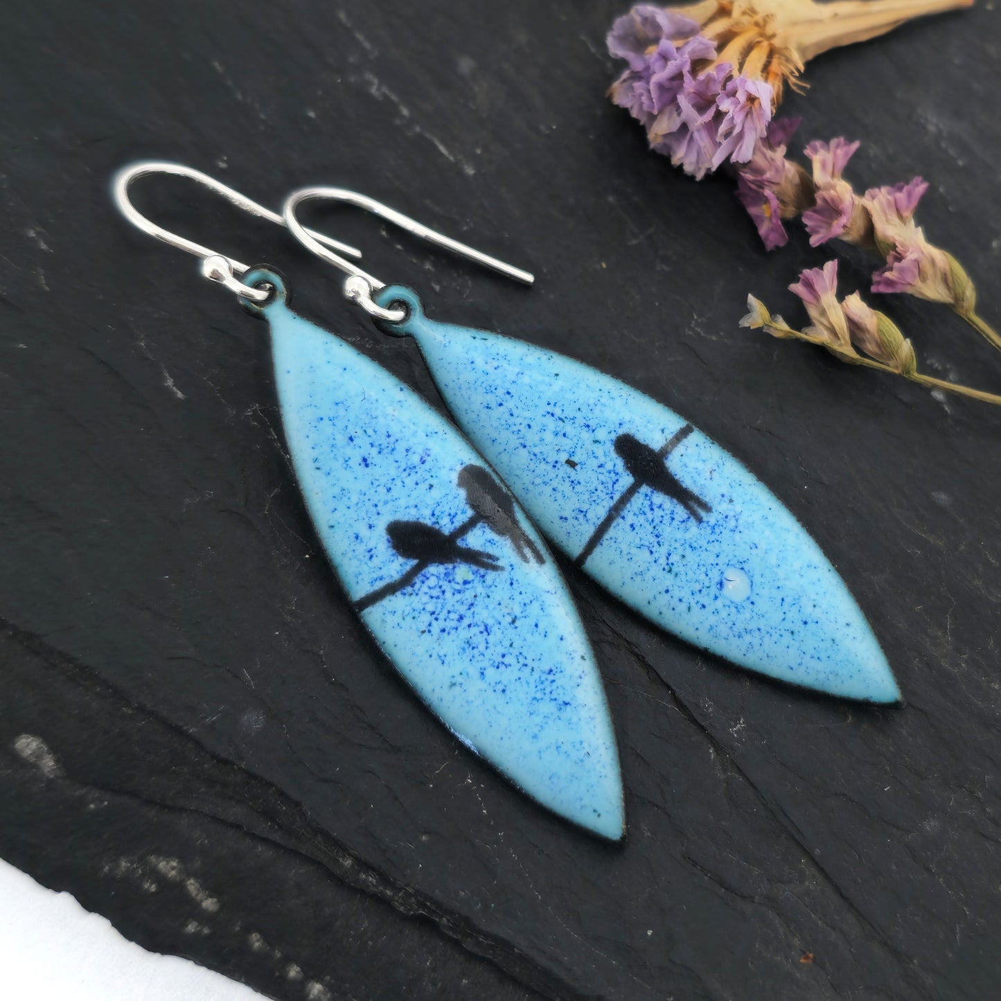 Boat-shaped enamel drop earrings with black coloured bird silhouettes sitting on a wire on a blue background. Pictured on slate with flowers.