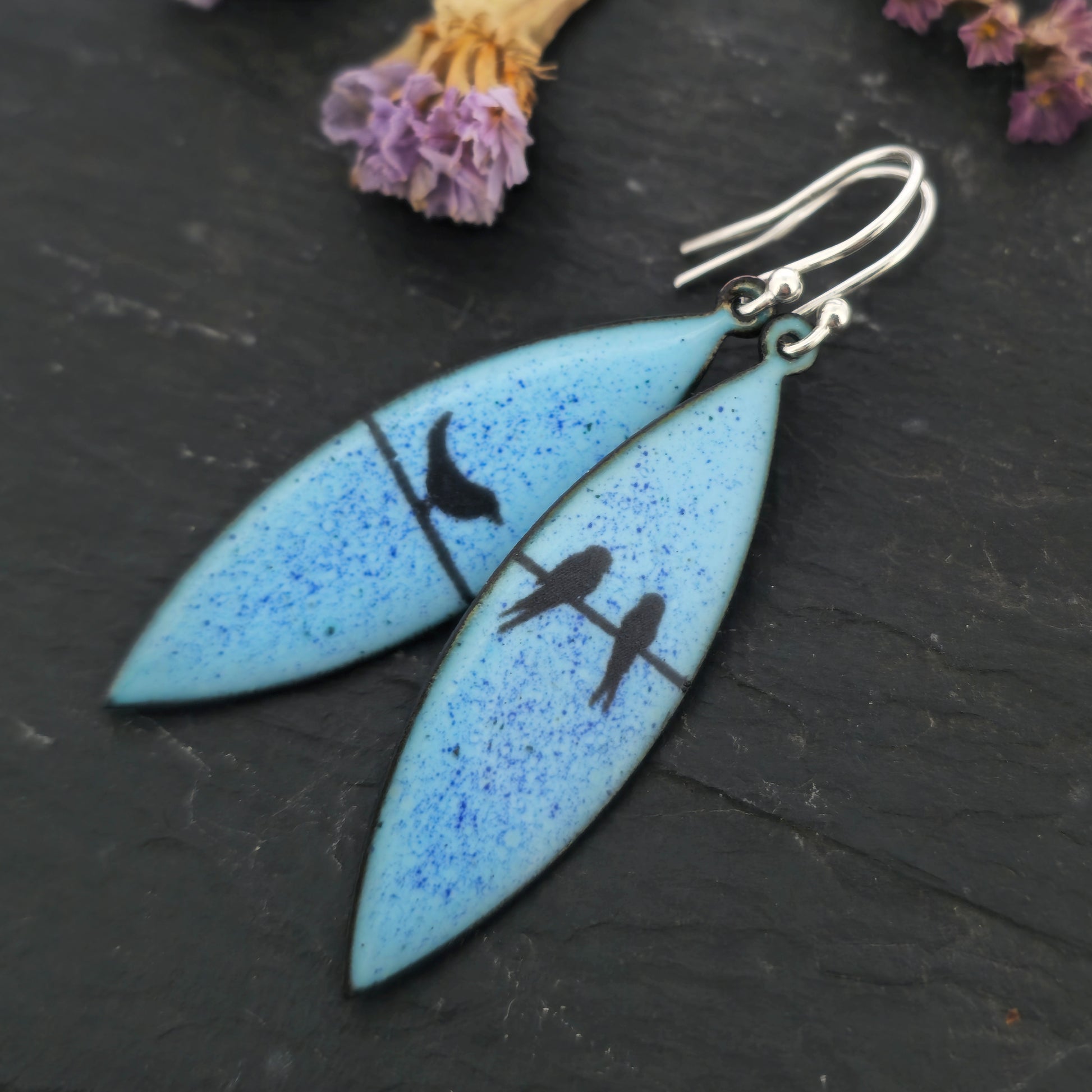 Boat-shaped enamel drop earrings with black coloured bird silhouettes sitting on a wire on a blue background. Pictured on slate with flowers.
