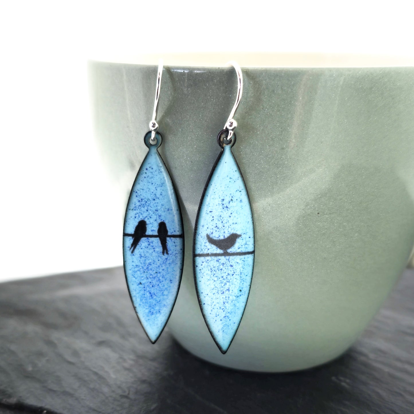 Boat-shaped enamel drop earrings with black coloured bird silhouettes sitting on a wire on a blue background. Pictured on a cup.