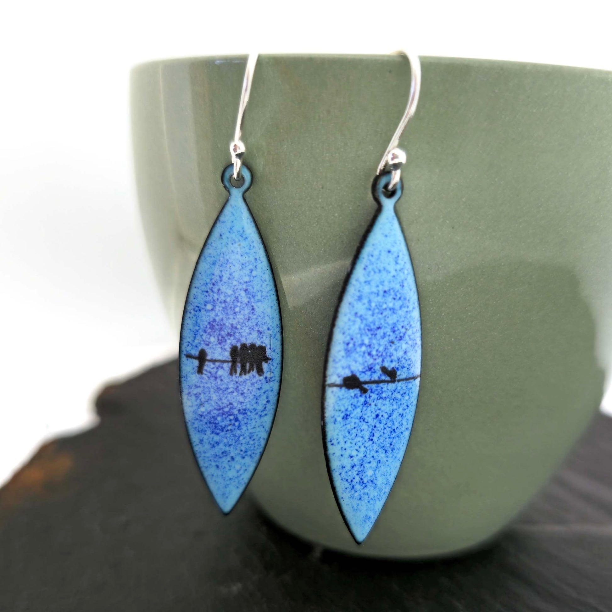 Boat-shaped enamel drop earrings with black bird silhouettes sitting on a wire on a blue background - pictured on a cup.