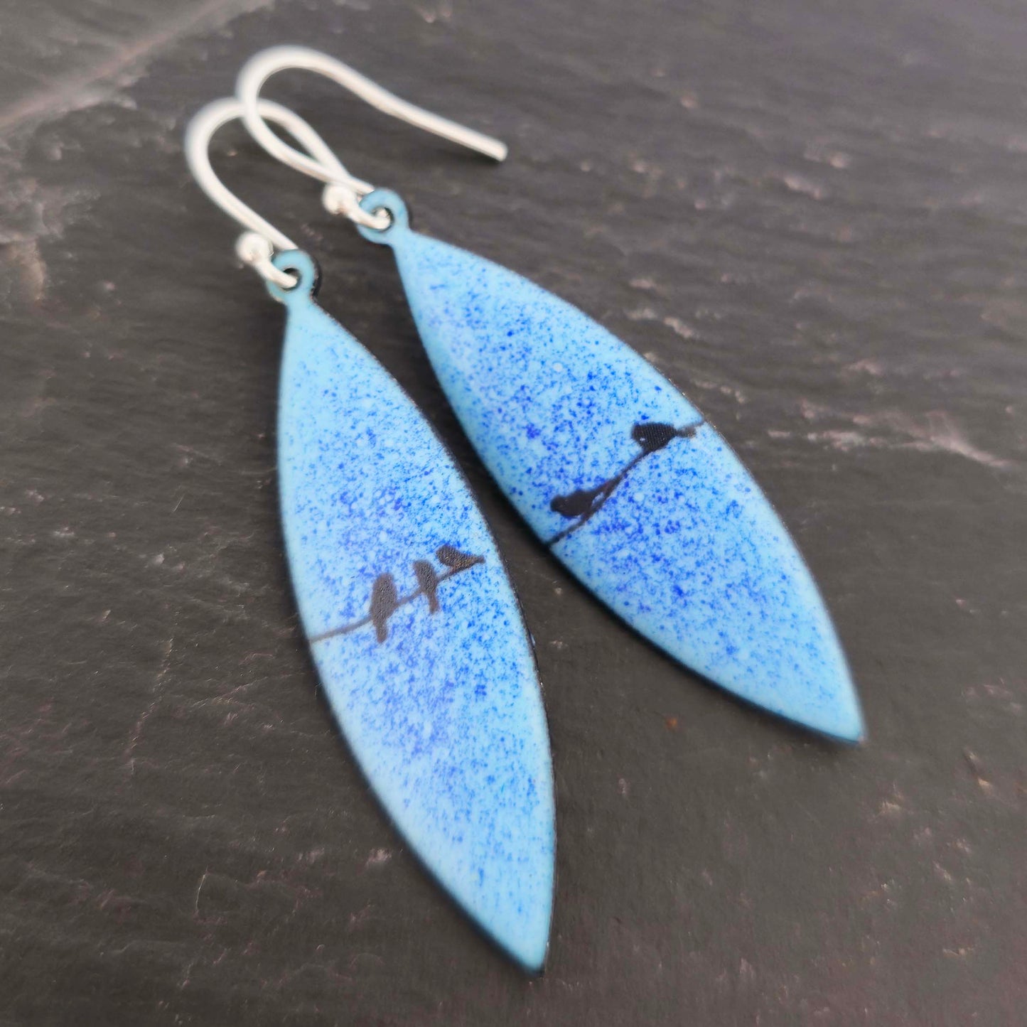Boat-shaped enamel drop earrings with black coloured bird silhouettes sitting on a wire on a blue background. Shown on slate.