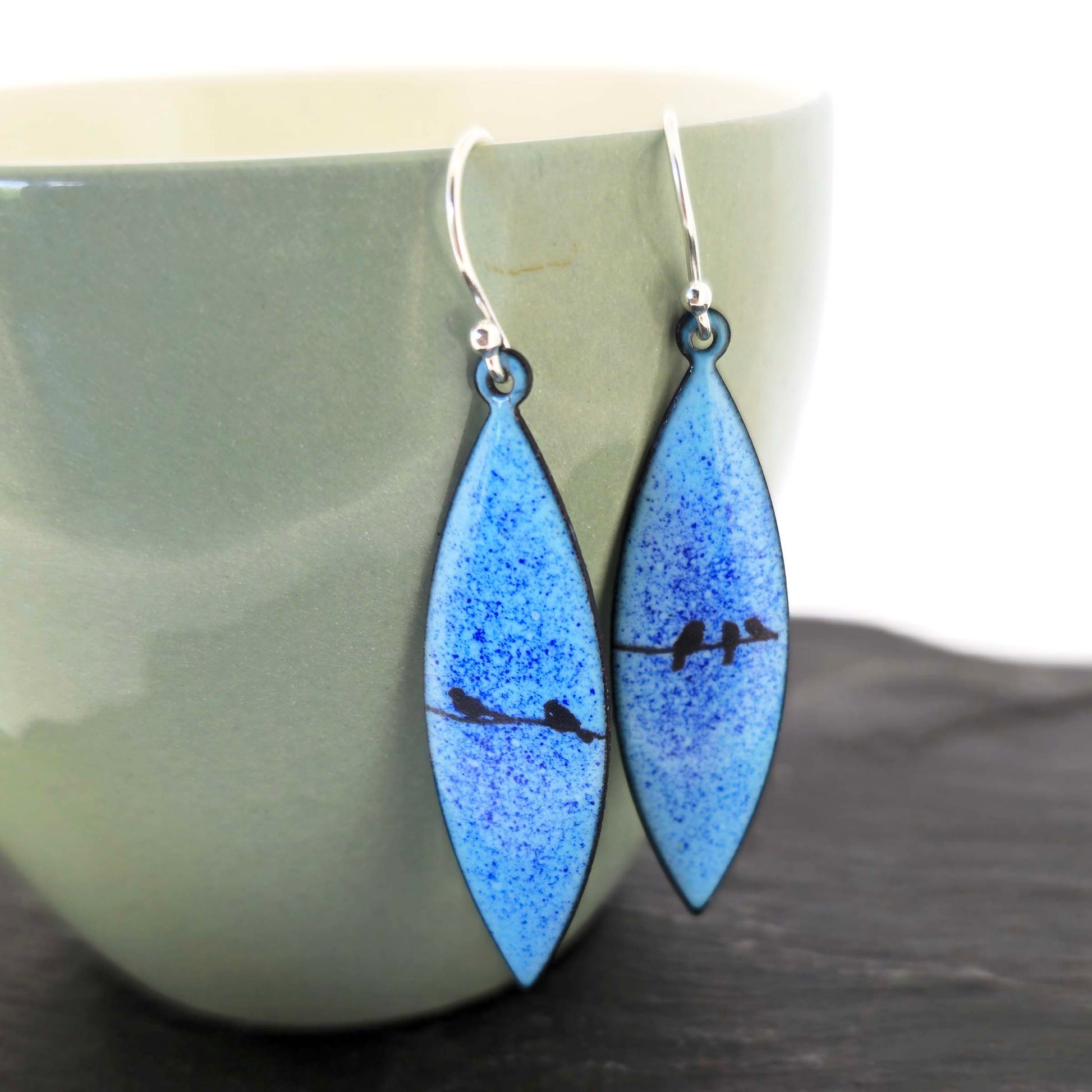 Boat-shaped enamel drop earrings with black coloured bird silhouettes sitting on a wire on a blue background. Shown on a green cup.