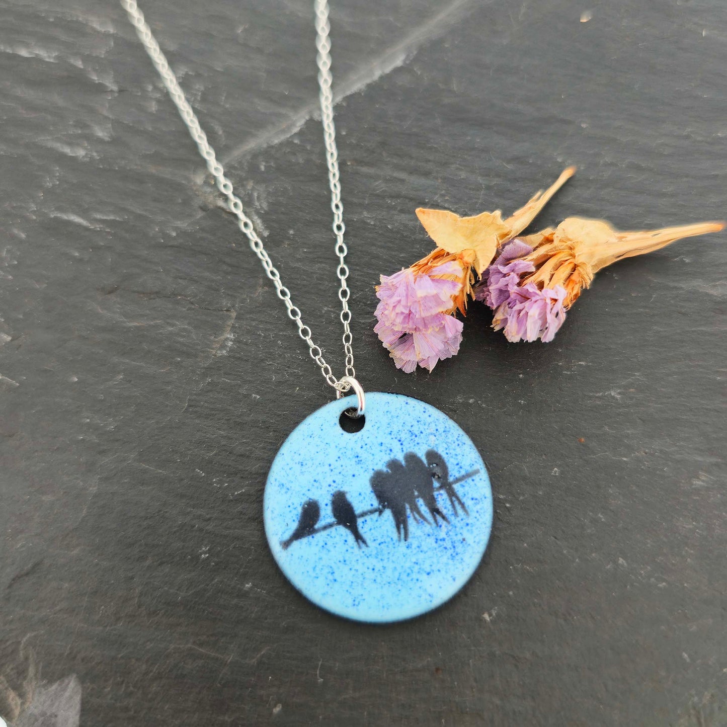Necklace with a blue enamel pendant featuring silhouettes of birds on a dark surface with dried flowers.