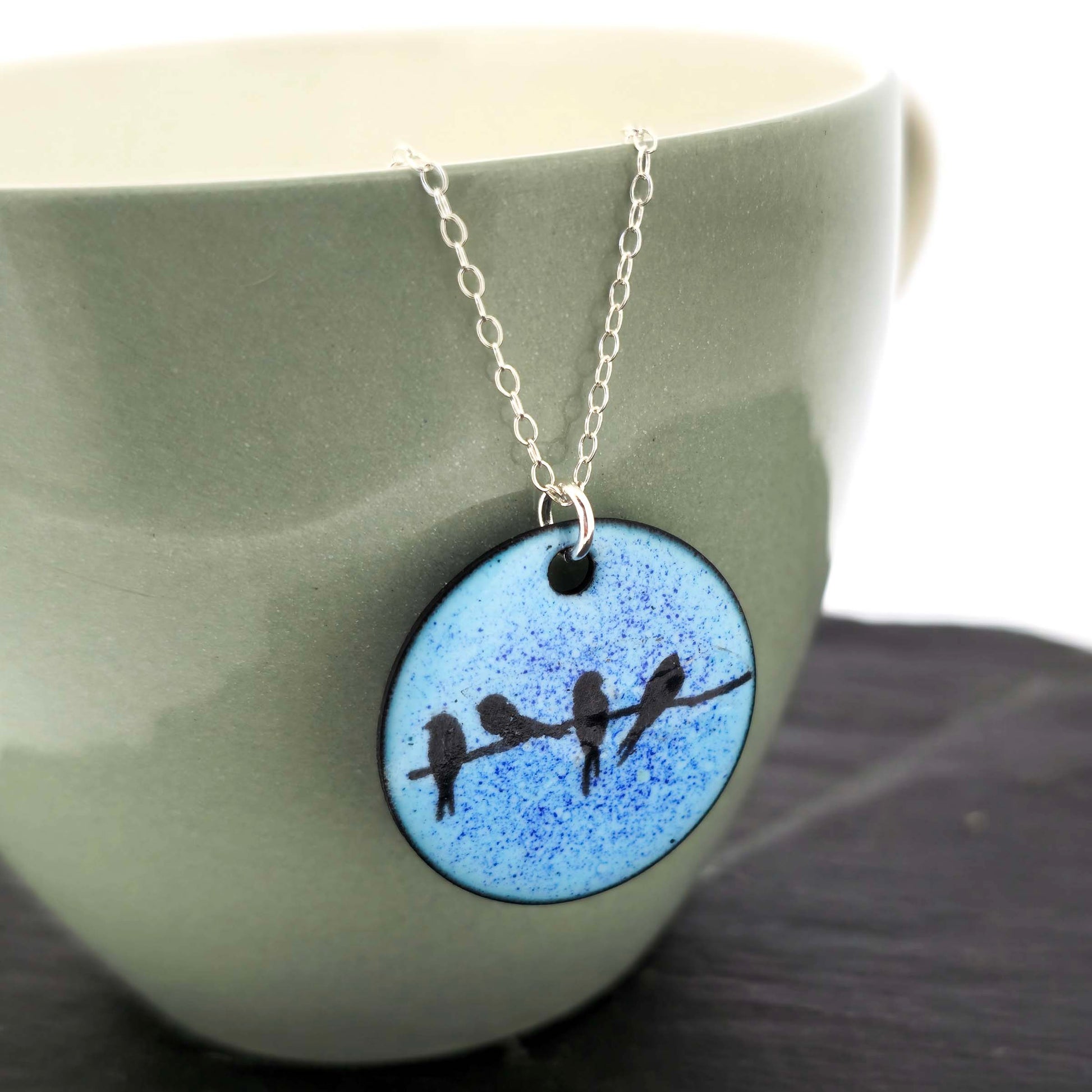 Necklace with a blue enamel pendant featuring silhouettes of birds displayed against a green ceramic background.