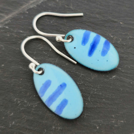 Pair of blue enamel oval earrings with a stripe pattern and silver ear hooks on a slate surface