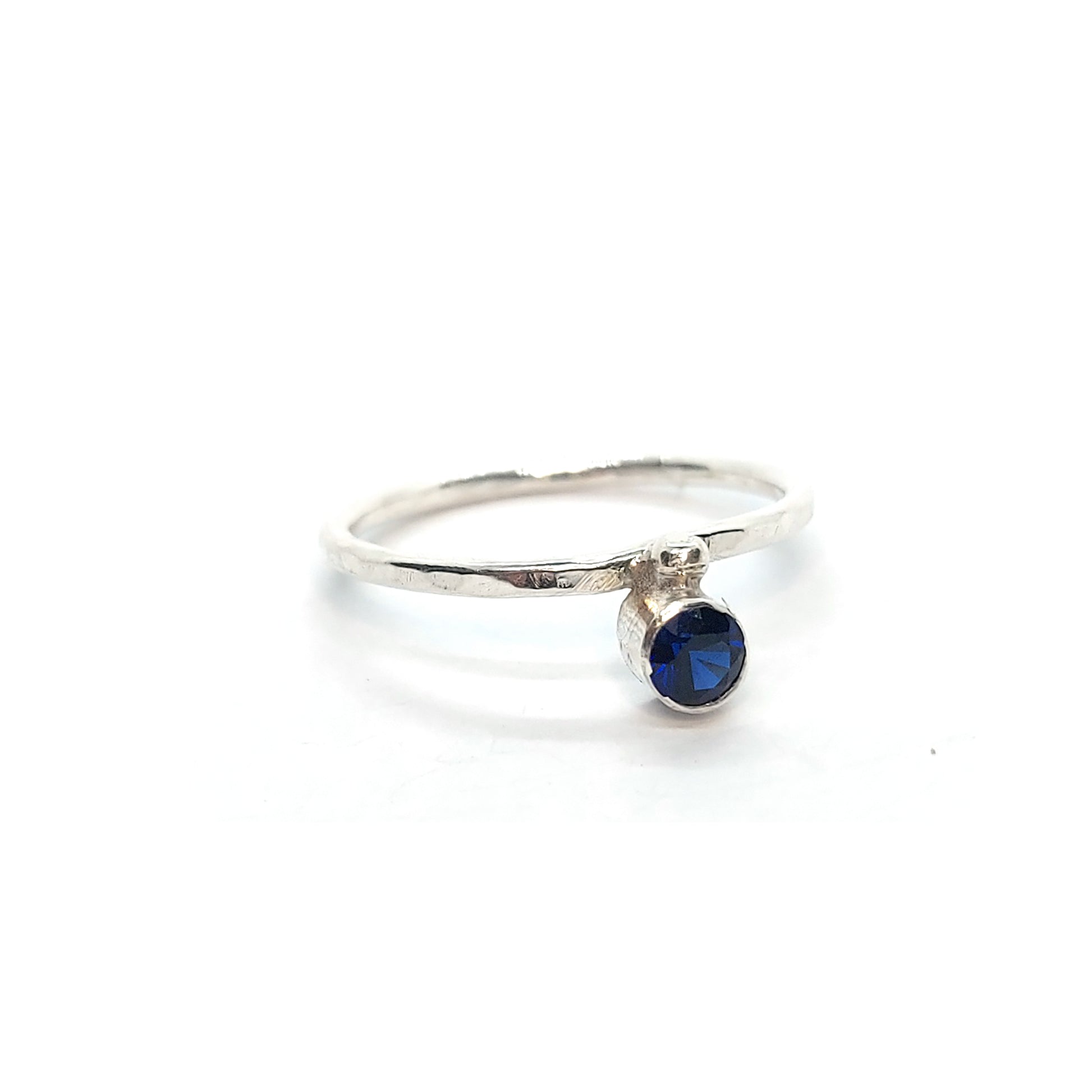 A silver thin ring with a hammered finish. The ring has a dark blue sapphire bezel set alongside a silver dot.
