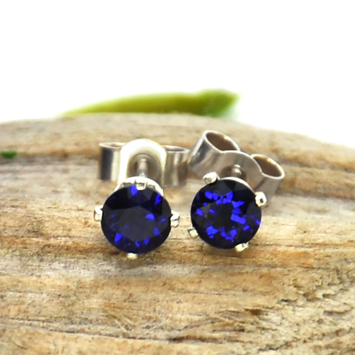 Silver 4 claw stud earrings set with dark blue sapphires. On wood. 4mm