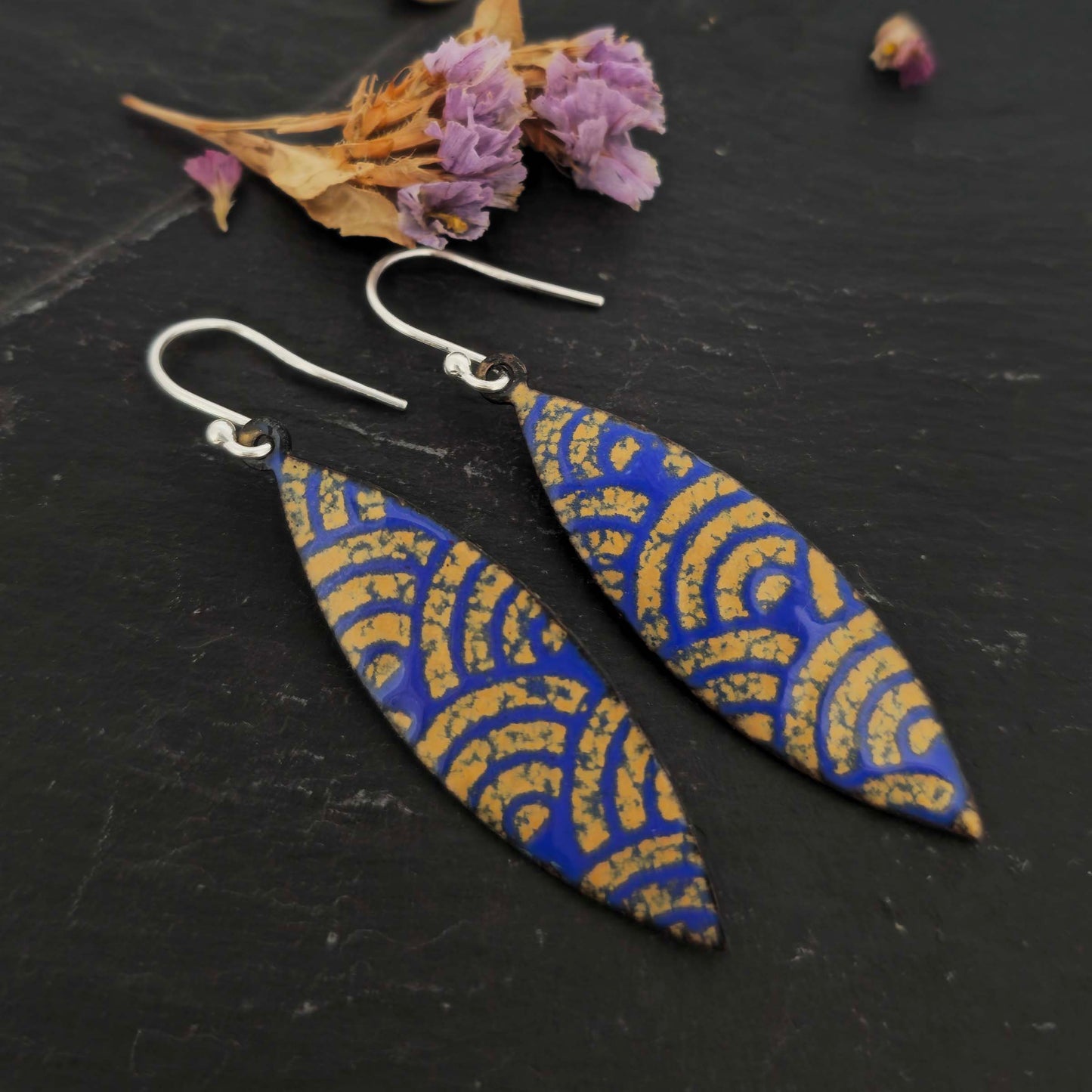 Pair of blue and gold enamel patterned earrings with silver ear hooks on dark slate with dried flowers