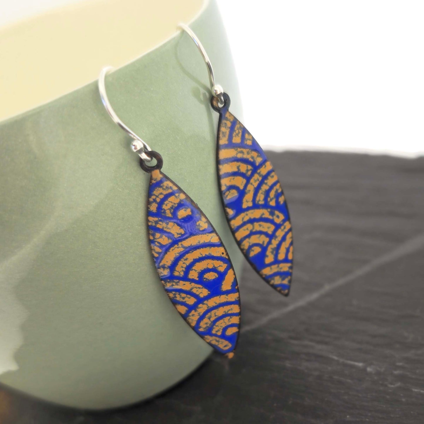 Pair of blue and gold enamel patterned earrings with silver ear hooks on a green cup