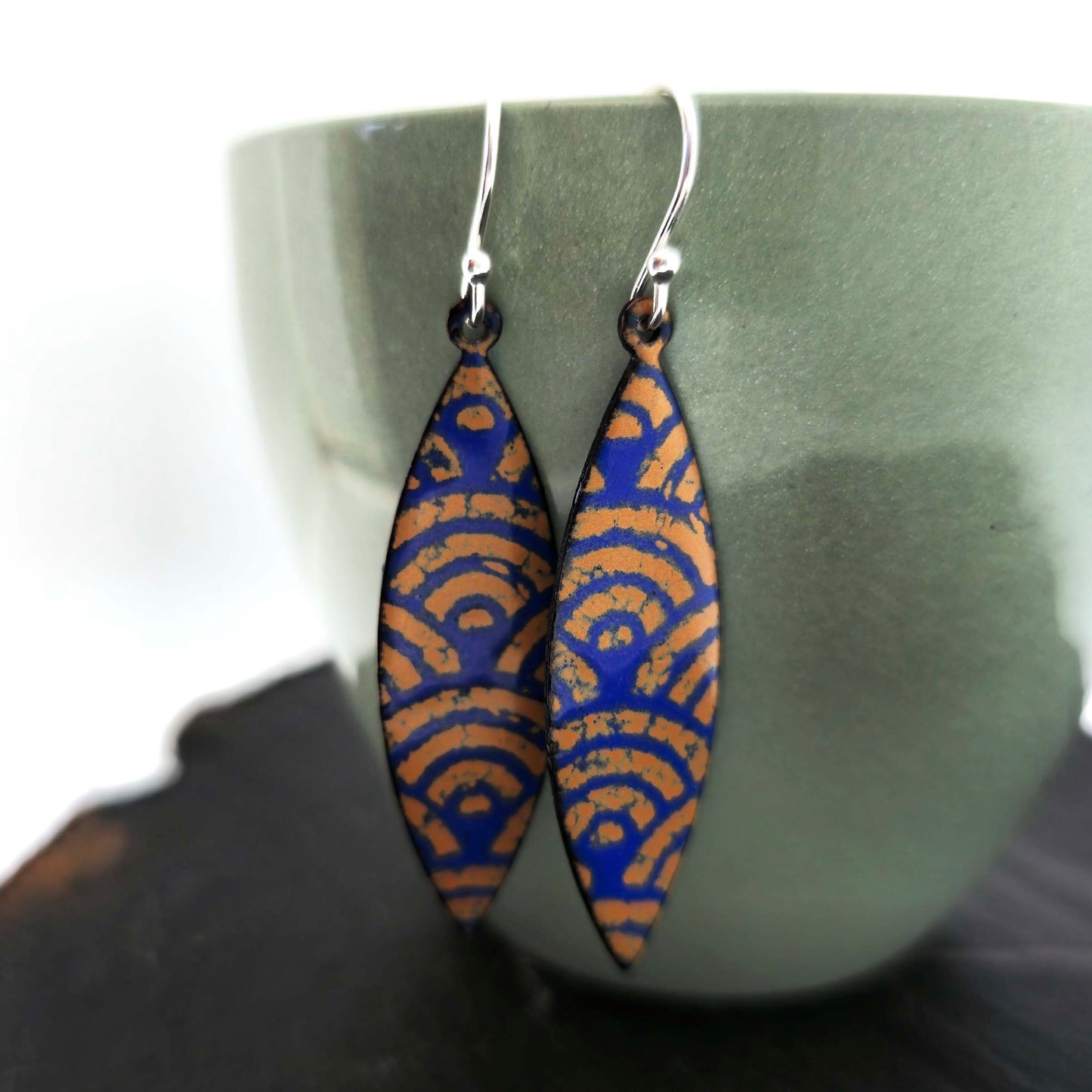 Pair of blue and gold enamel patterned earrings with silver ear hooks on a green cup