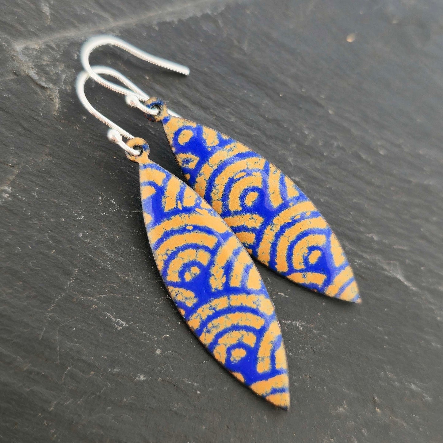 Pair of blue and gold enamel patterned earrings with silver ear hooks on a dark surface.