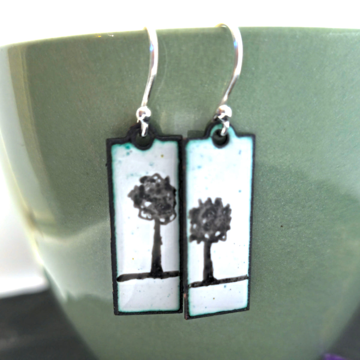 Rectangle enamel earrings with a single tree silhouette on each earring, in white, green and black, suspended from silver hooks. On a pale green background. Handmade. Mijoux Jewellery.