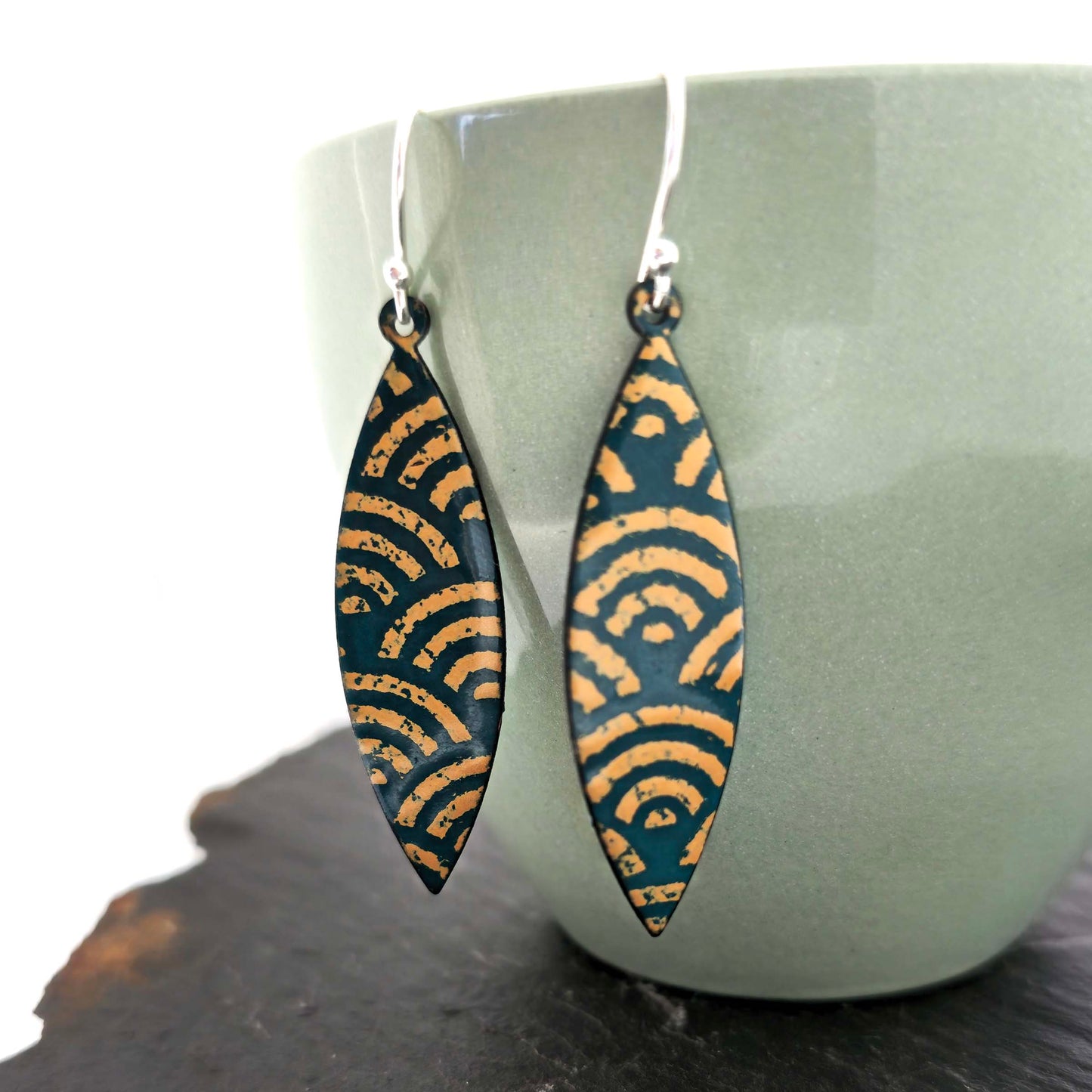 A pair of boat shaped drop earrings with a dark green background and an art deco style fan pattern in gold enamel. On silver ear hooks. Shown on a green cup.