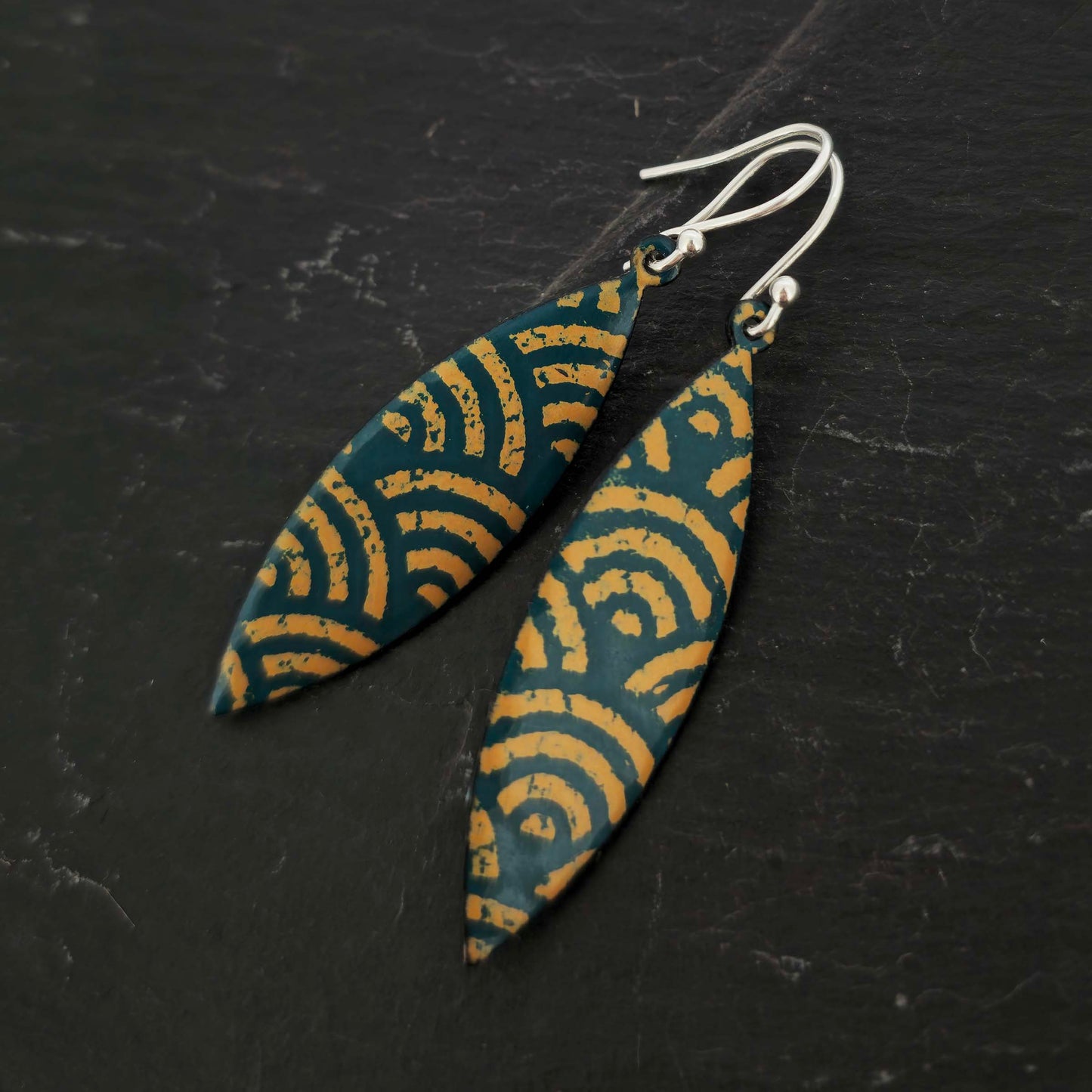 A pair of boat shaped drop earrings with a dark green background and an art deco style fan pattern in gold enamel. On silver ear hooks. Shown on slate.