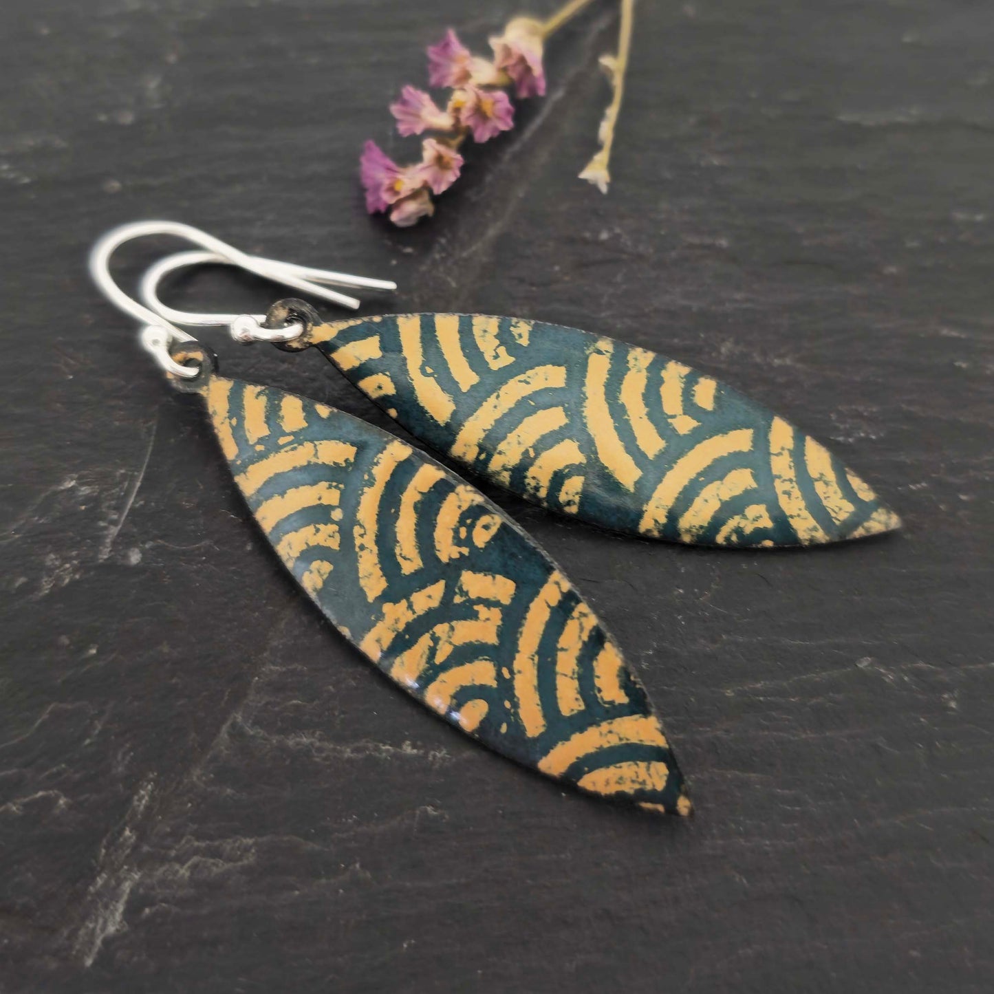 A pair of boat shaped drop earrings with a dark green background and an art deco style fan pattern in gold enamel. On silver ear hooks. Shown on slate with flowers.