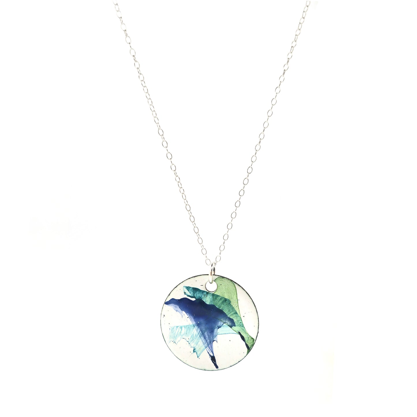 A round enamel pendant with a white background and splashes of dark blue, dark green and jade green. Suspended from a silver chain.