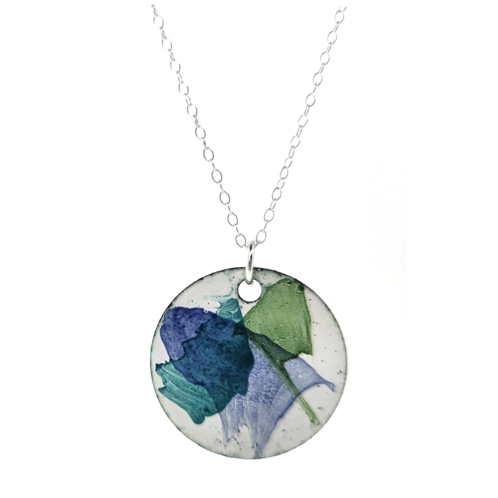 A round enamel pendant with a white background and splashes of dark blue, dark green and jade green. Suspended from a silver chain. Mijoux Creations.