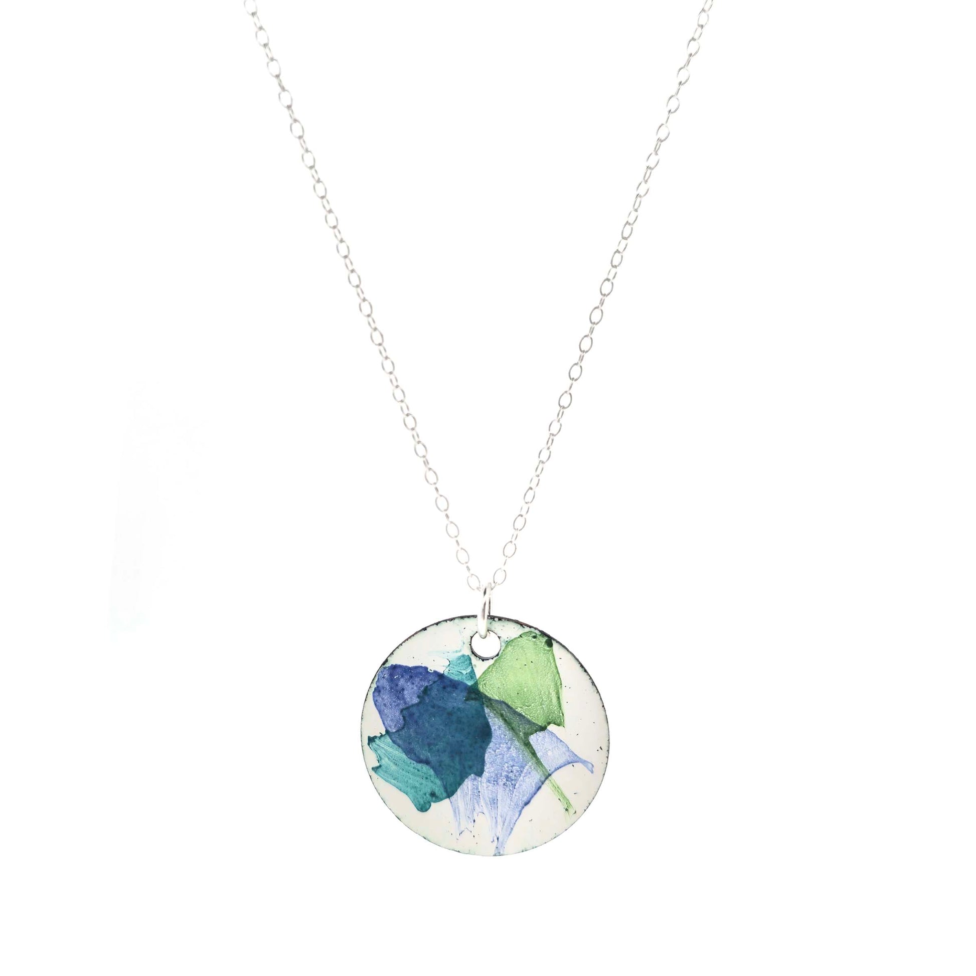 A round enamel pendant with a white background and splashes of dark blue, dark green and jade green. Suspended from a silver chain. Mijoux Creations