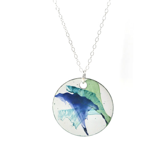 A round enamel pendant with a white background and splashes of dark blue, dark green and jade green. Suspended from a silver chain.