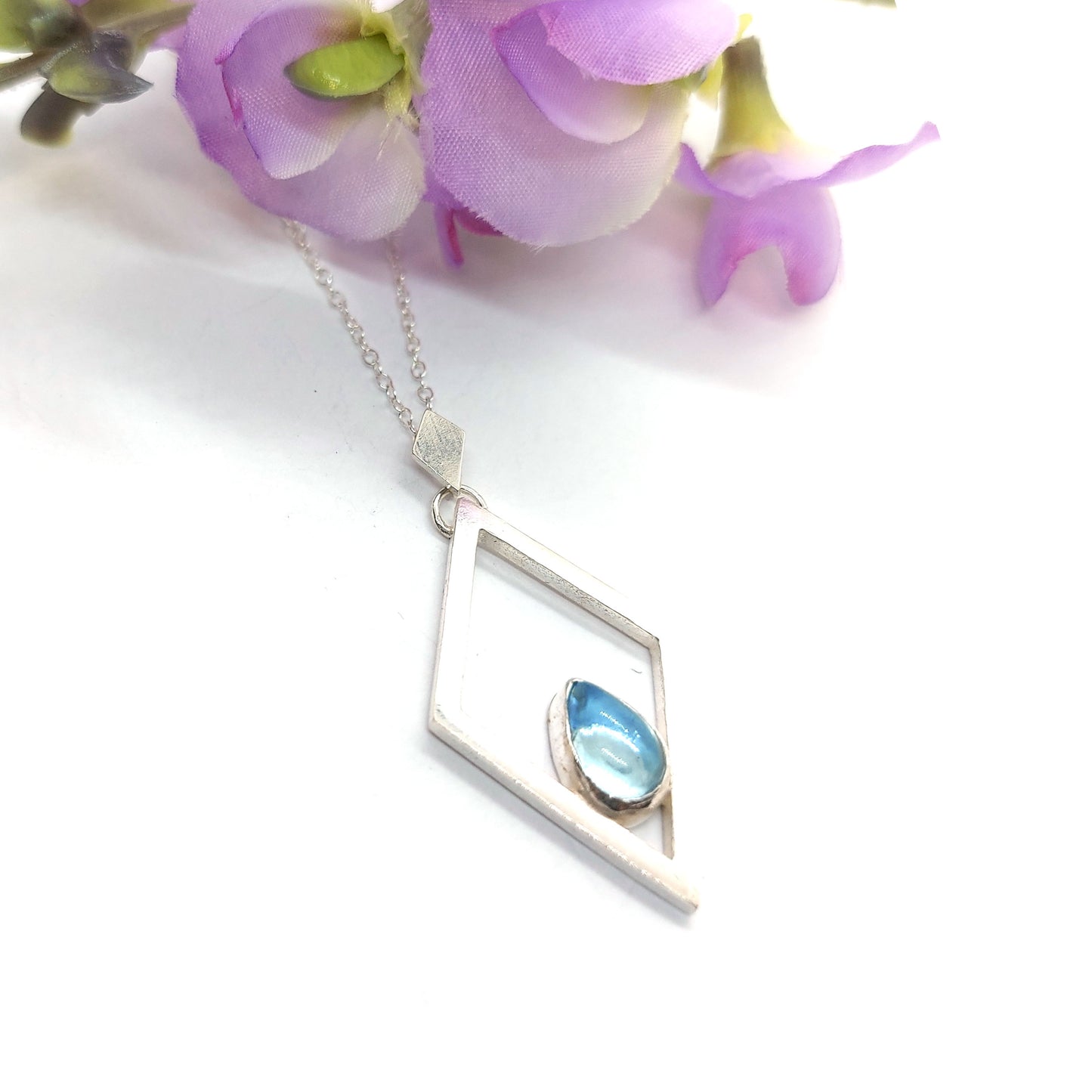 A silver open diamond shaped pendant with a teardrop shaped blue topaz gemstone set inside. A small diamond shaped bail attaches the pendant to a silver chain. Pictured with flowers.