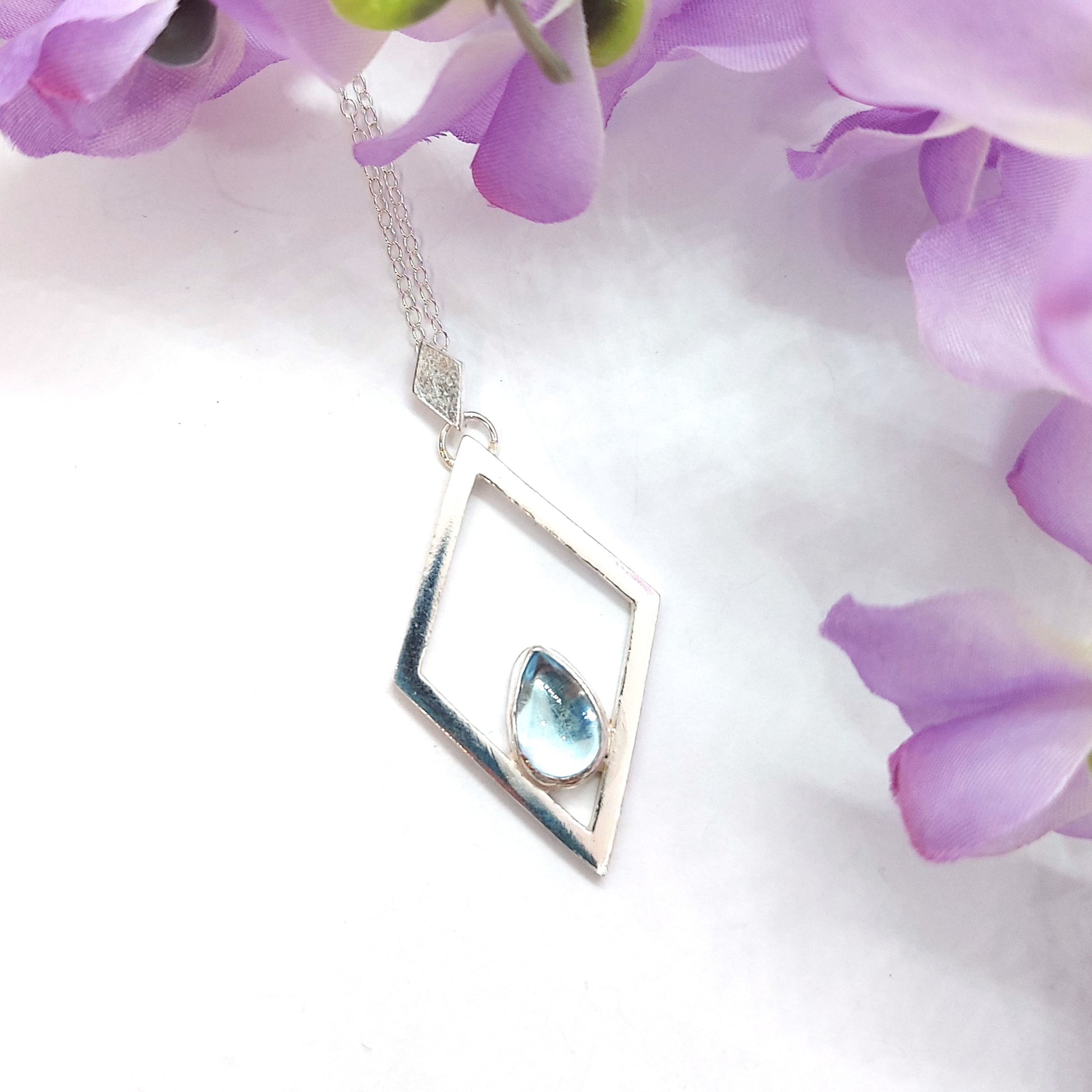 A silver open diamond shaped pendant with a teardrop shaped blue topaz gemstone set inside. A small diamond shaped bail attaches the pendant to a silver chain. Pictured with flowers.