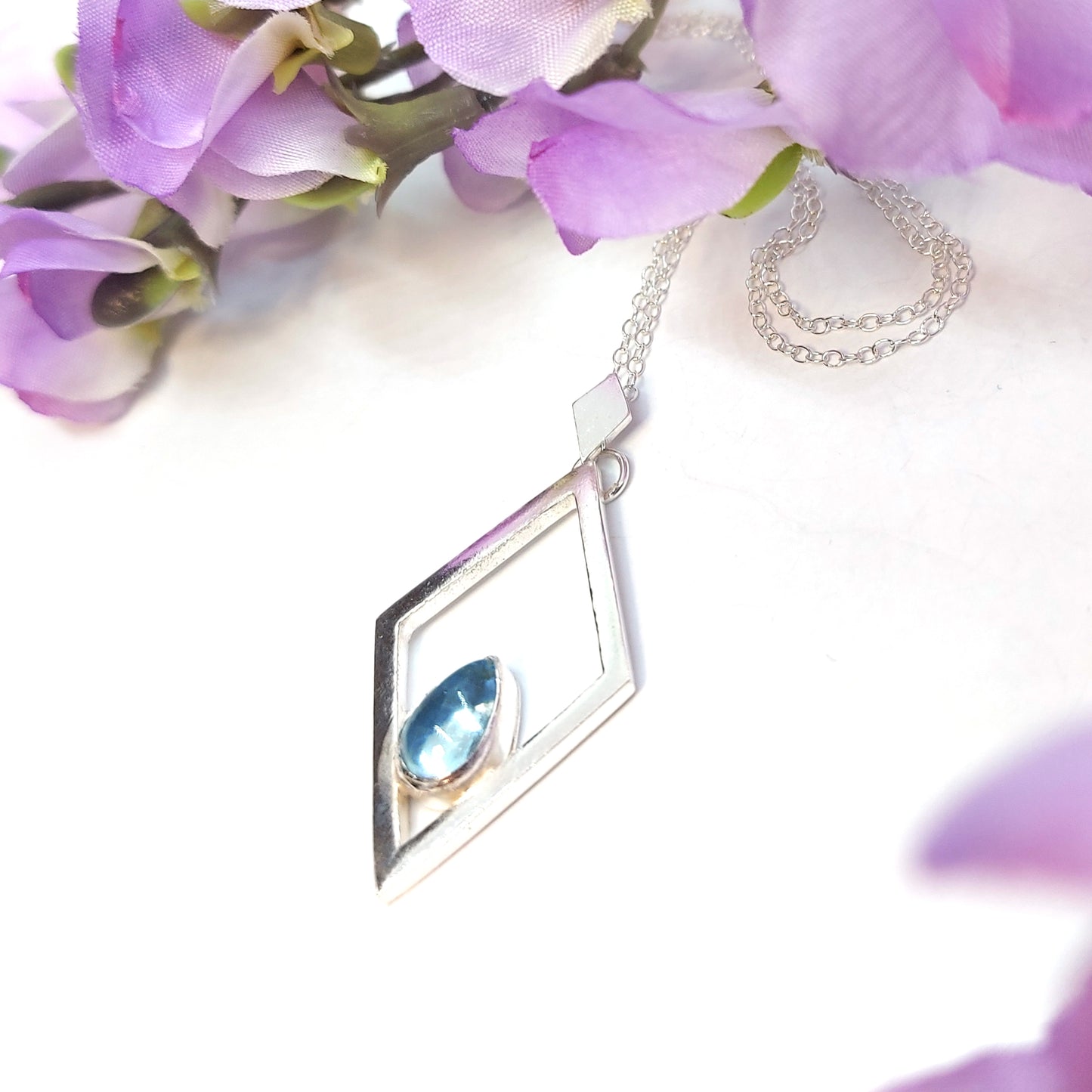 A silver open diamond shaped pendant with a teardrop shaped blue topaz gemstone set inside. A small diamond shaped bail attaches the pendant to a silver chain. Pictured with flowers.