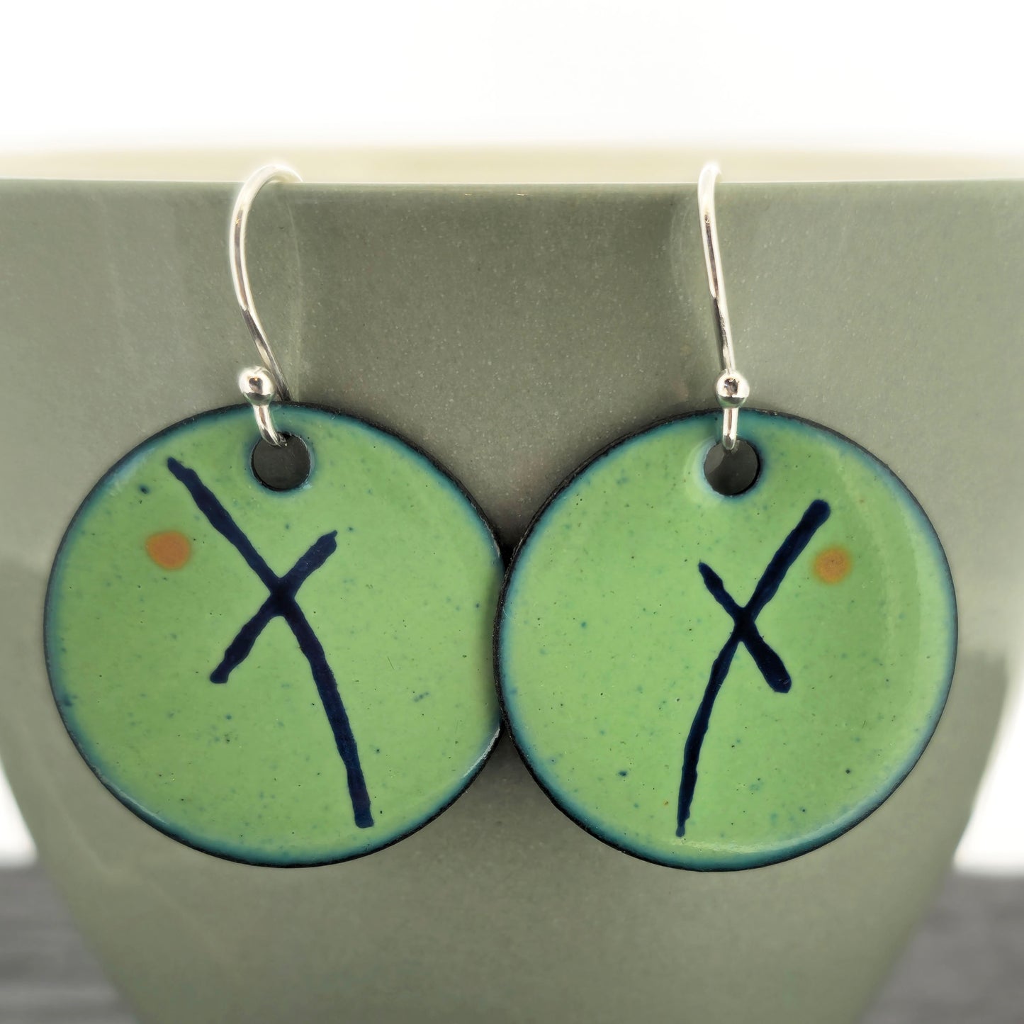 A pair of handmade round green enamel drop earrings with an asymmetric blue cross and orange dot. On sterling silver ear hooks. On a pale green cup. Mijoux jewellery.