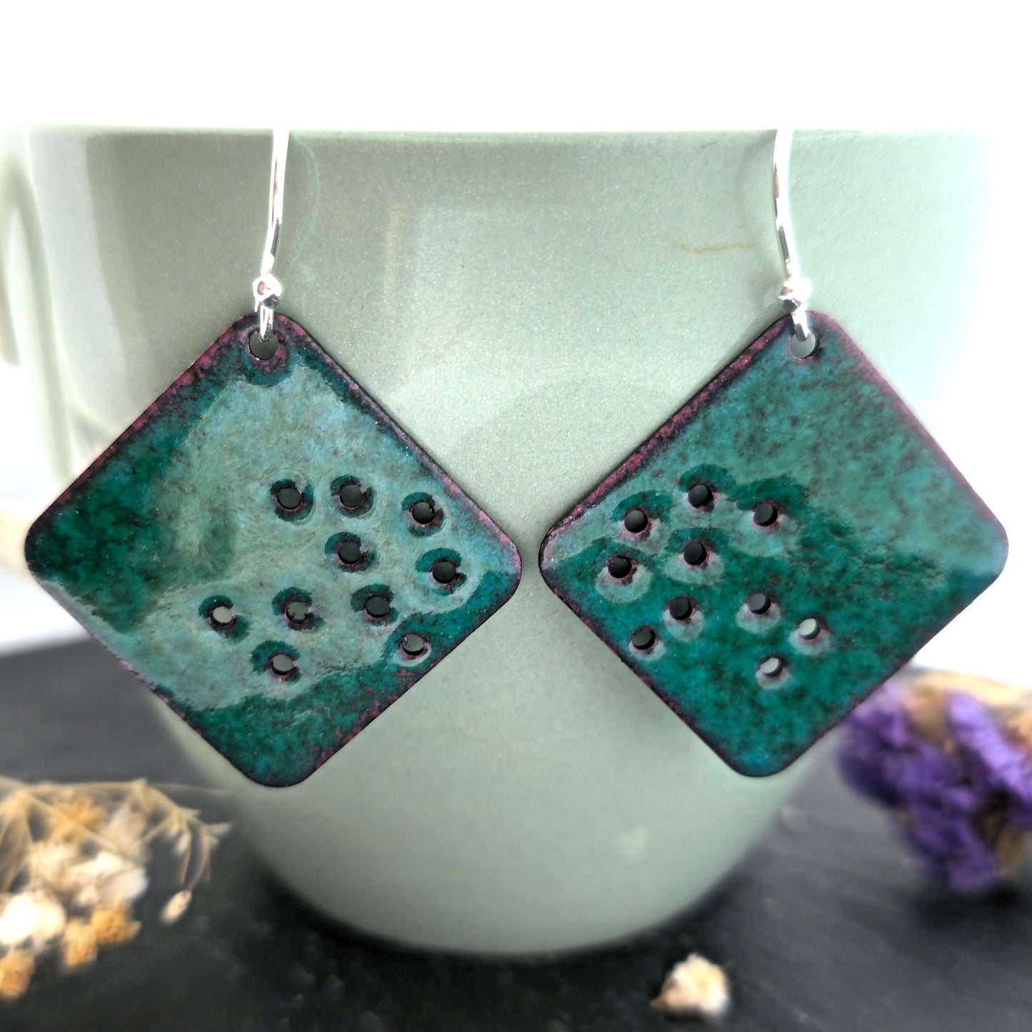 Pair of square-shaped earrings with a speckled green enamel surface and small holes, on a light green cup background. Handmade. Mijoux Jewellery