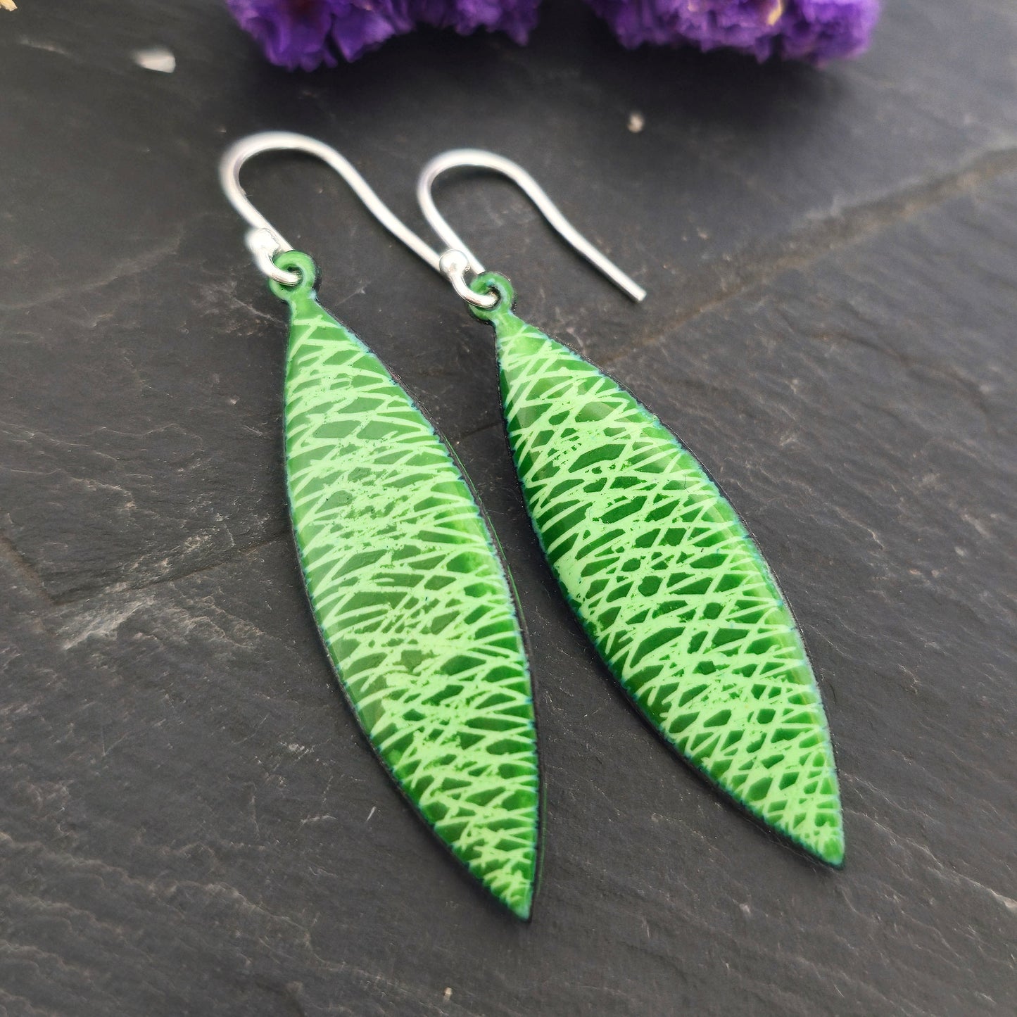 Handmade boat-shaped drop earrings with a green enamel scatter line pattern, suspended from silver ear hooks. Shown on a black background with flowers. Mijoux Jewellery.