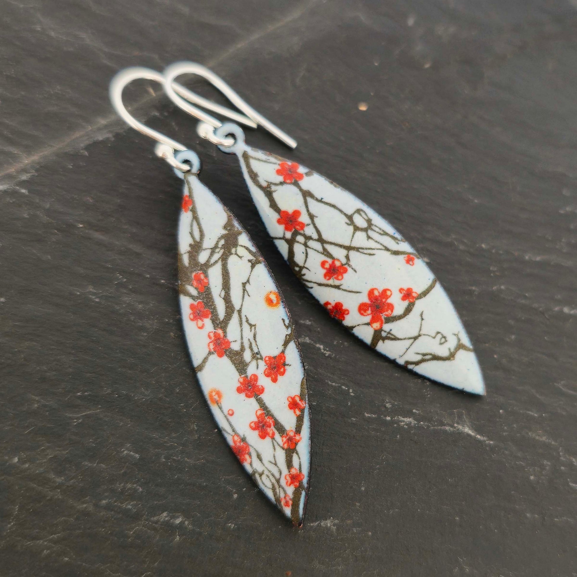 Boat-shaped enamel drop earrings with branches of red cherry blossom on a grey background.