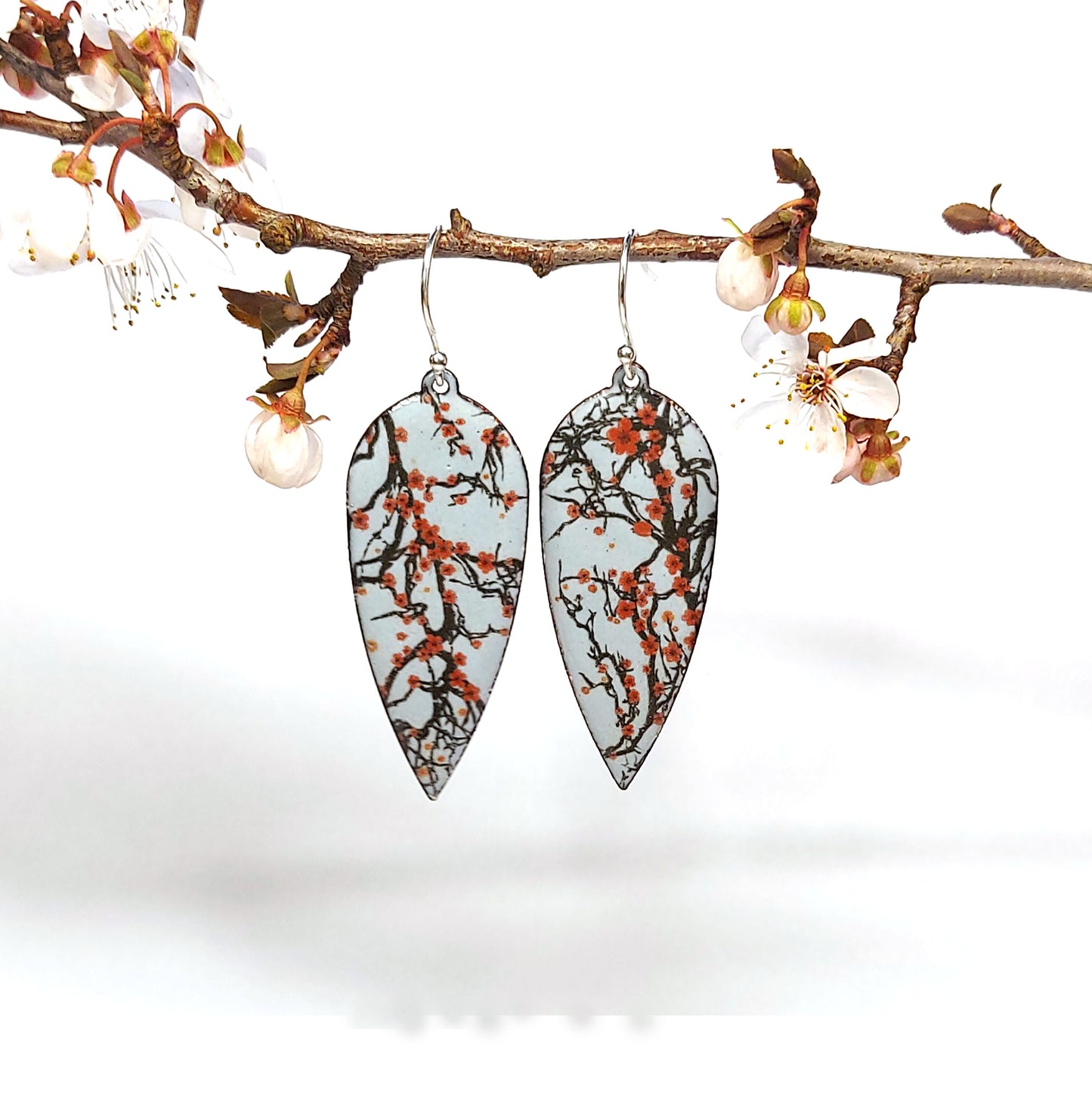 Statement enamel earrings featuring branches and red cherry blossom on a grey enamel background. On silver ear hooks. Suspended from a twig.