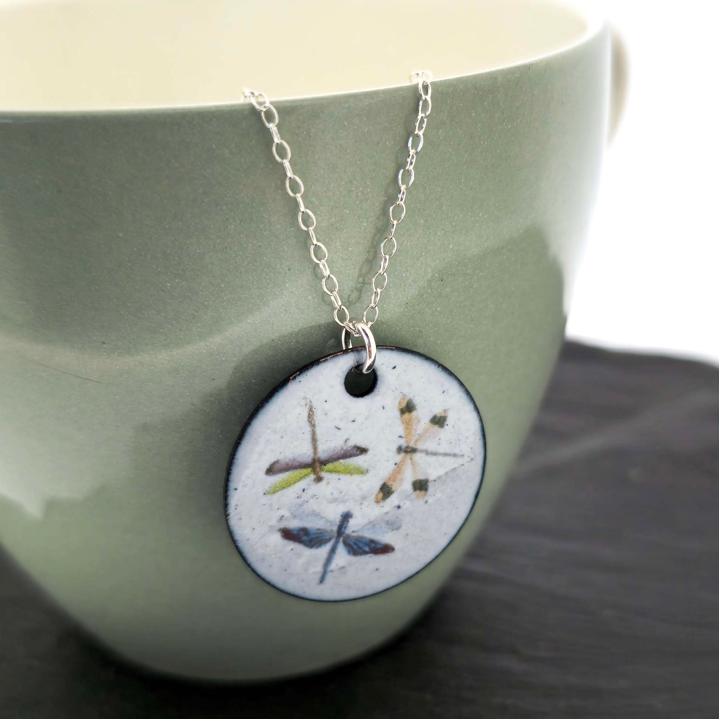 Necklace with a round enamel pendant featuring three dragonfly designs on a silver chain. Shown on a ceramic cup.