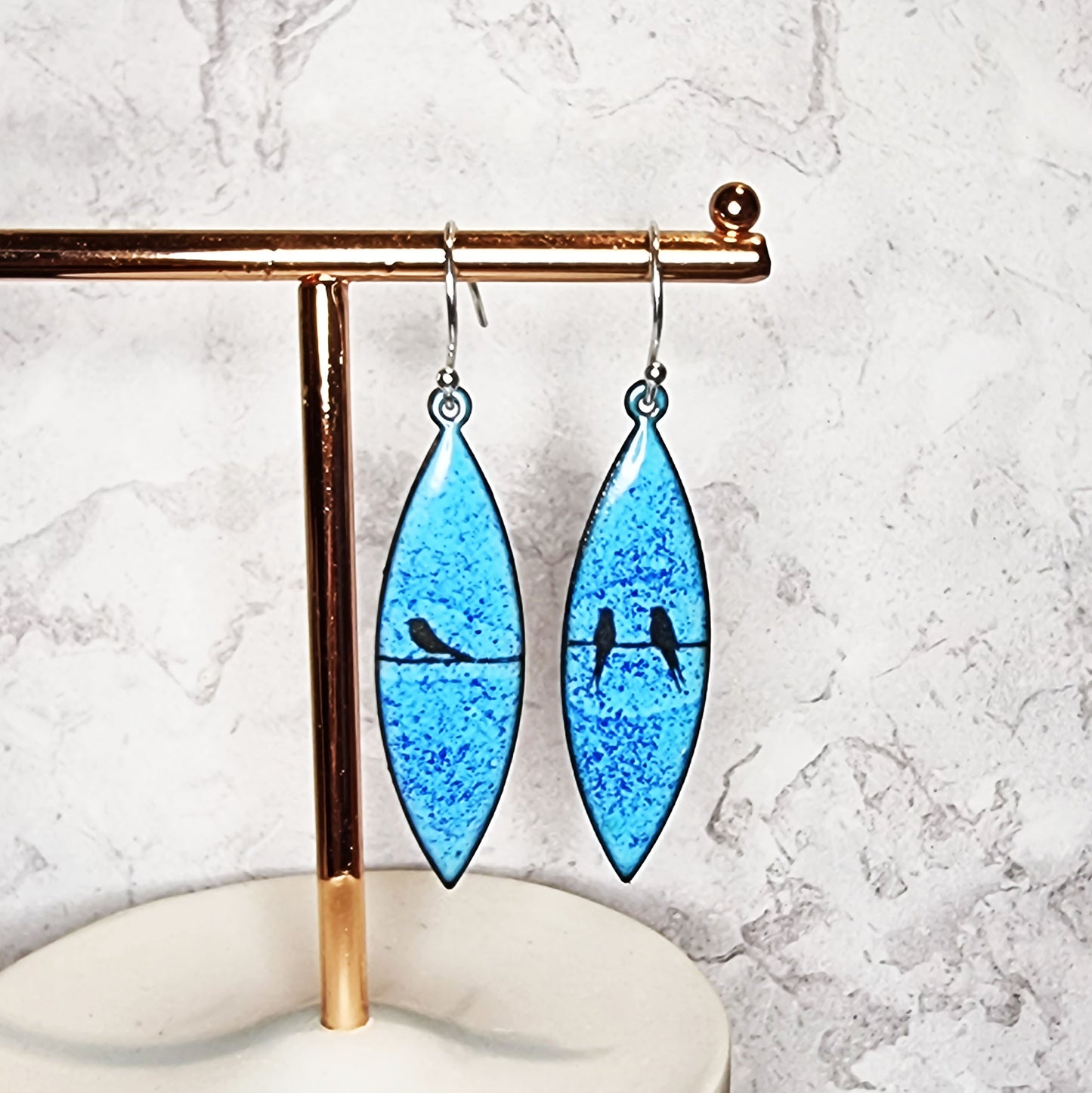 Boat-shaped enamel drop earrings with black coloured bird silhouettes sitting on a wire on a blue background - on stand