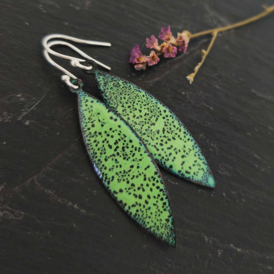 A pair of boat shaped drop earrings with a speckled dark and light green enamel pattern. On silver ear hooks. Shown on slate with flowers.