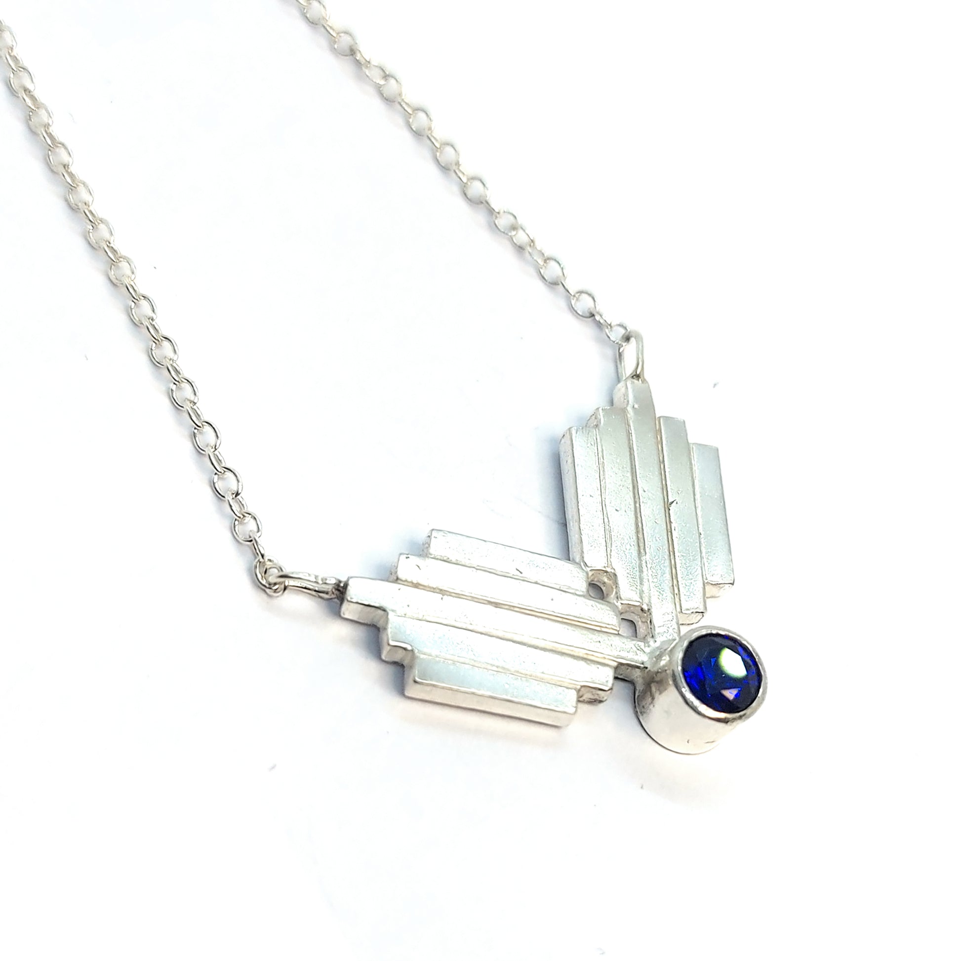 A silver Art Deco style necklace with a focal point of 2 lined elements and a dark blue sapphire gemstone.