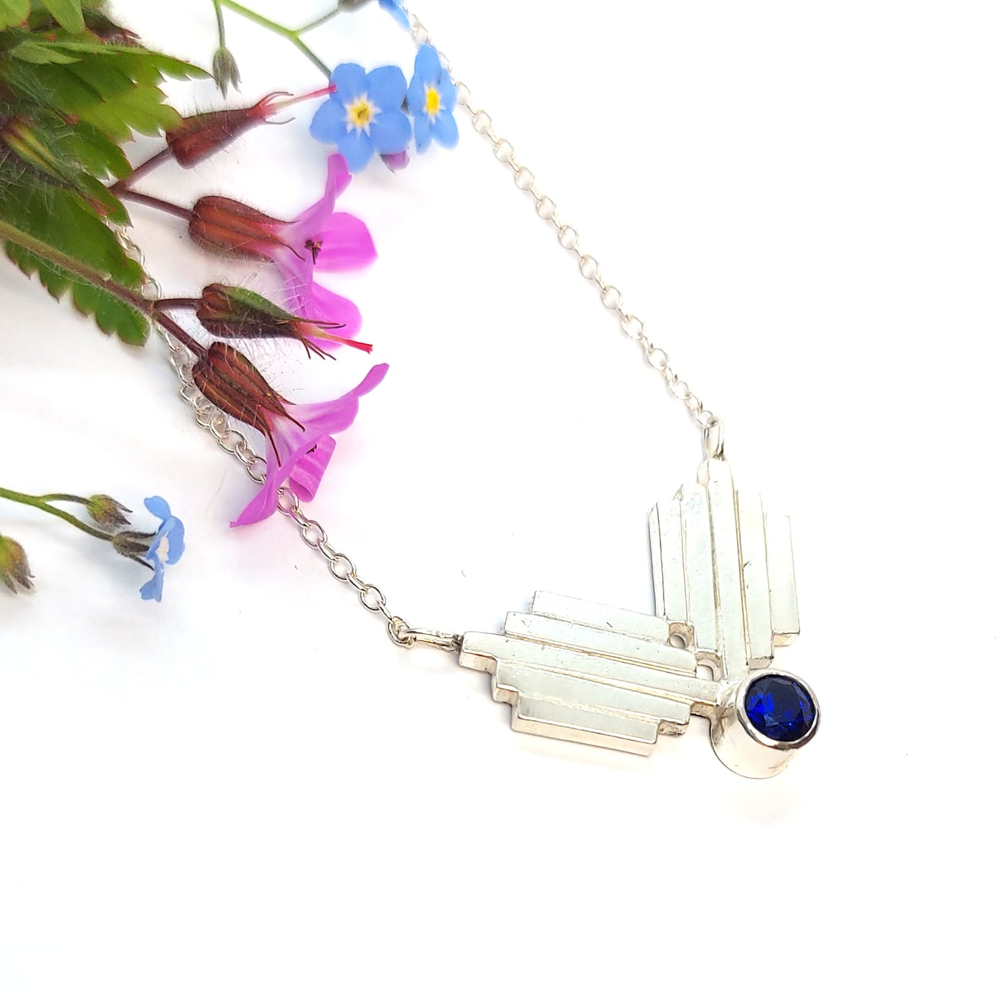 A silver Art Deco style necklace with a focal point of 2 lined elements and a dark blue sapphire gemstone. Pictured with flowers.