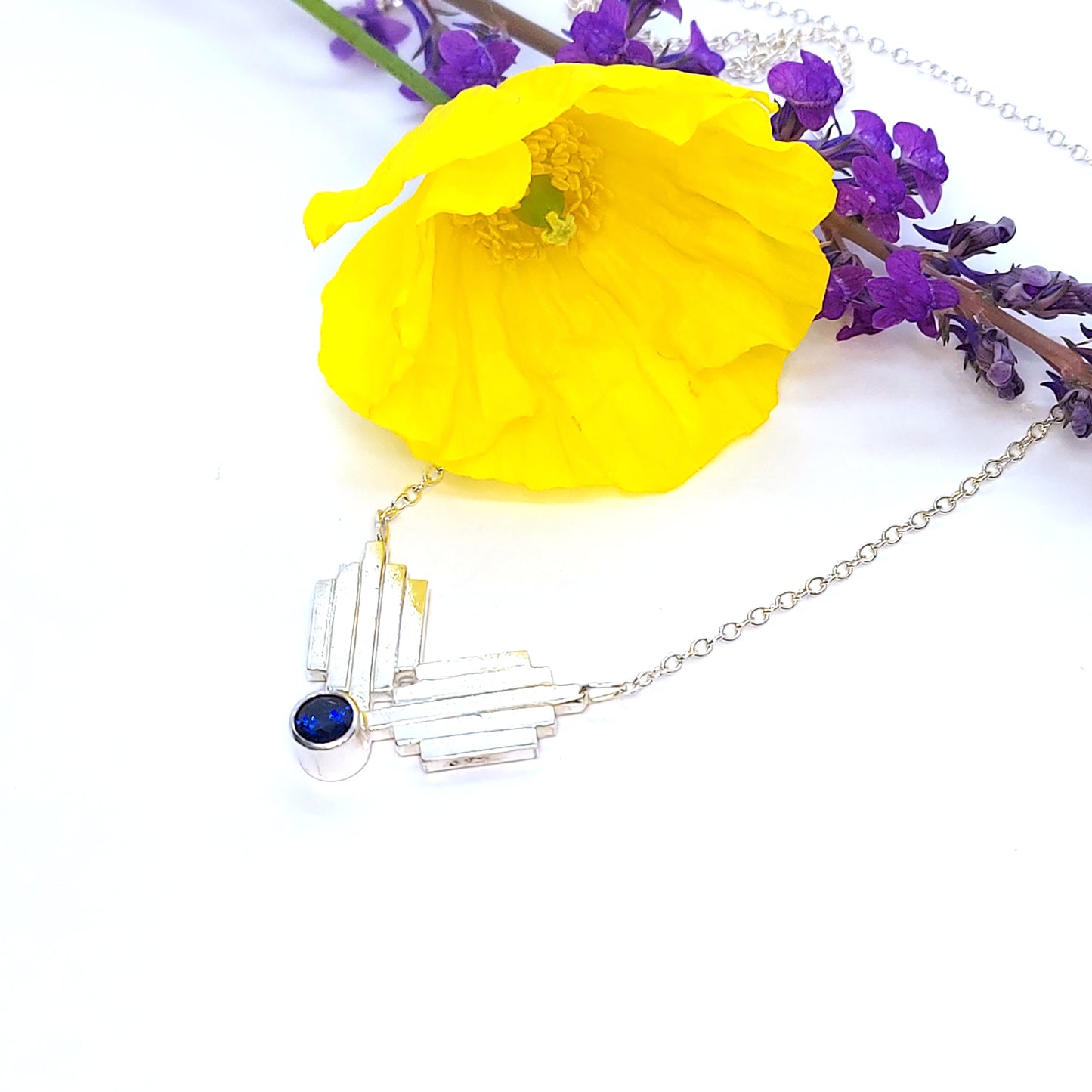 A silver Art Deco style necklace with a focal point of 2 lined elements and a dark blue sapphire gemstone. Pictured with flowers.