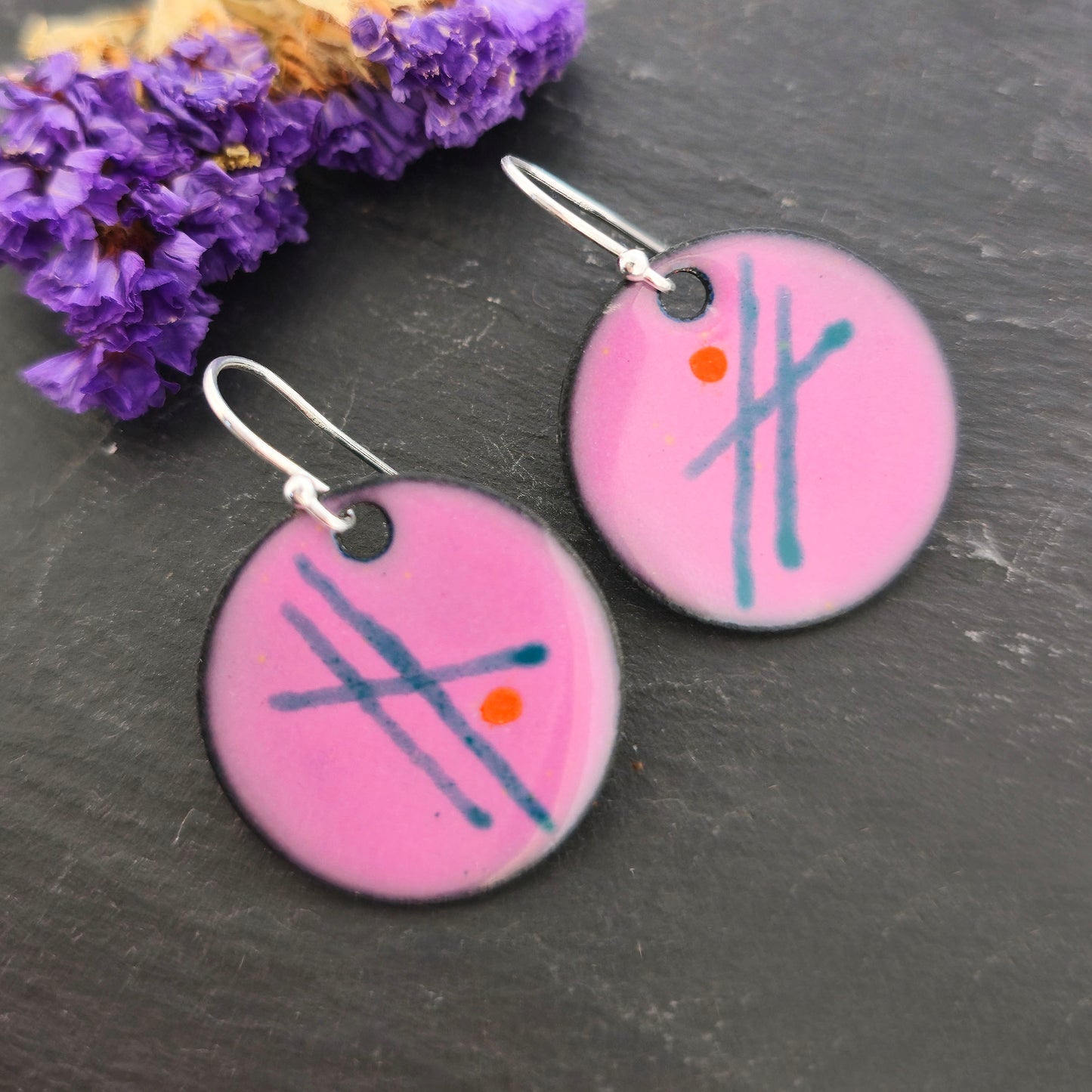 A pair of round pink enamel drop earrings with green lines and orange dots. On silver ear hooks. On a black background with purple flowers. Mijoux jewellery.